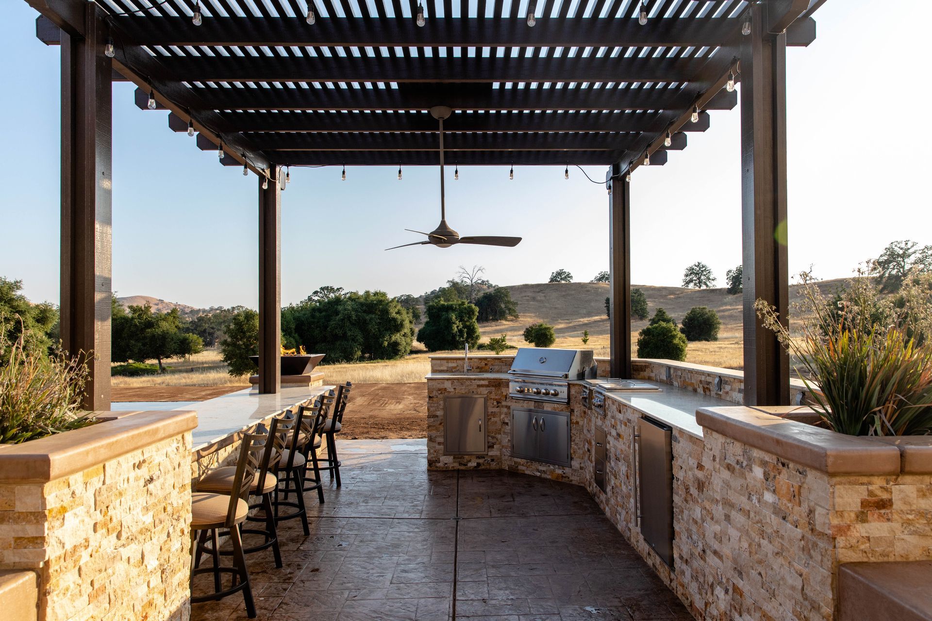 Covered outdoor patio with bar seating, stone counters, and a ceiling fan overlooking a sunny landscape