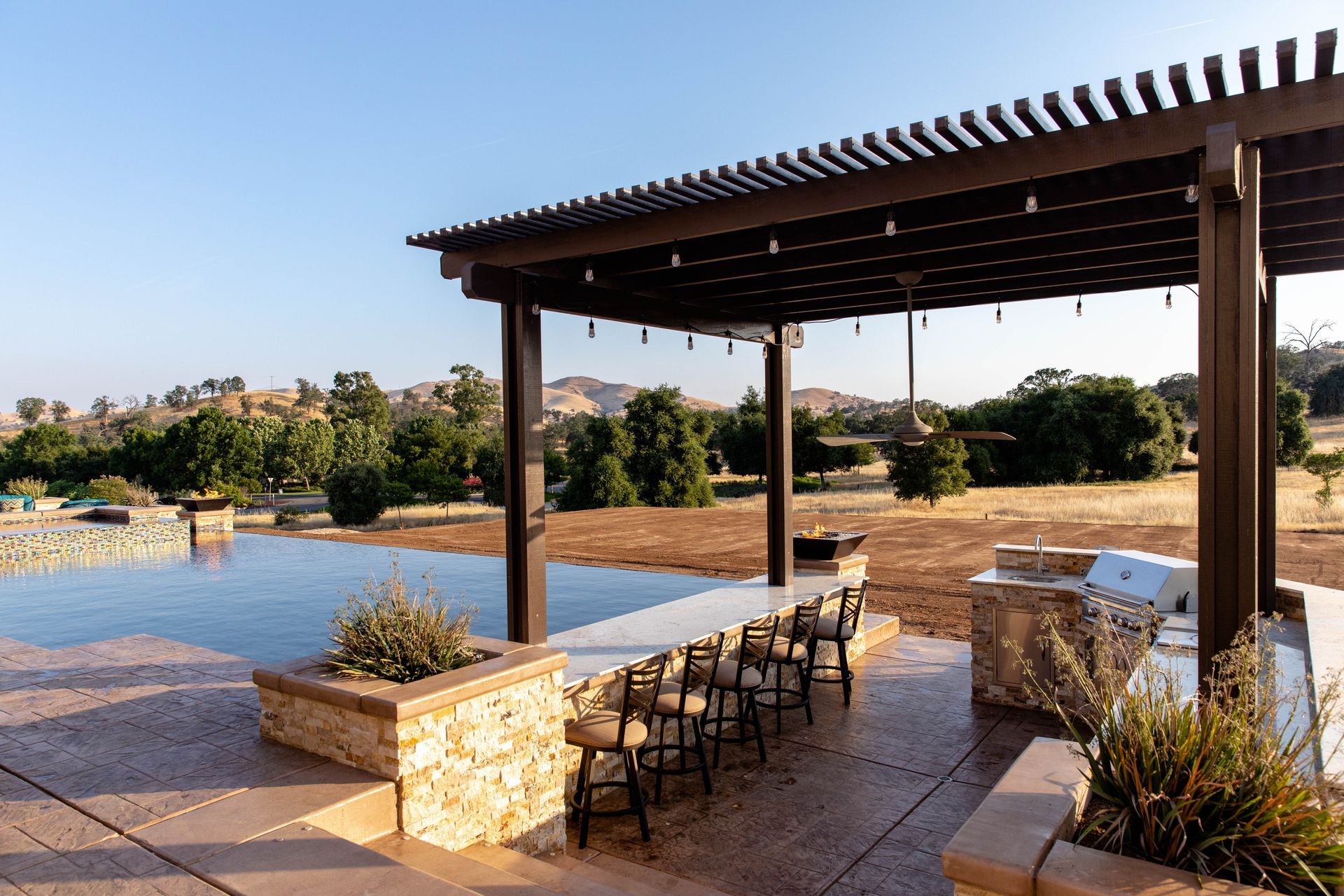 Outdoor patio with stone seating, bar stools, and a pergola beside a pool at sunset