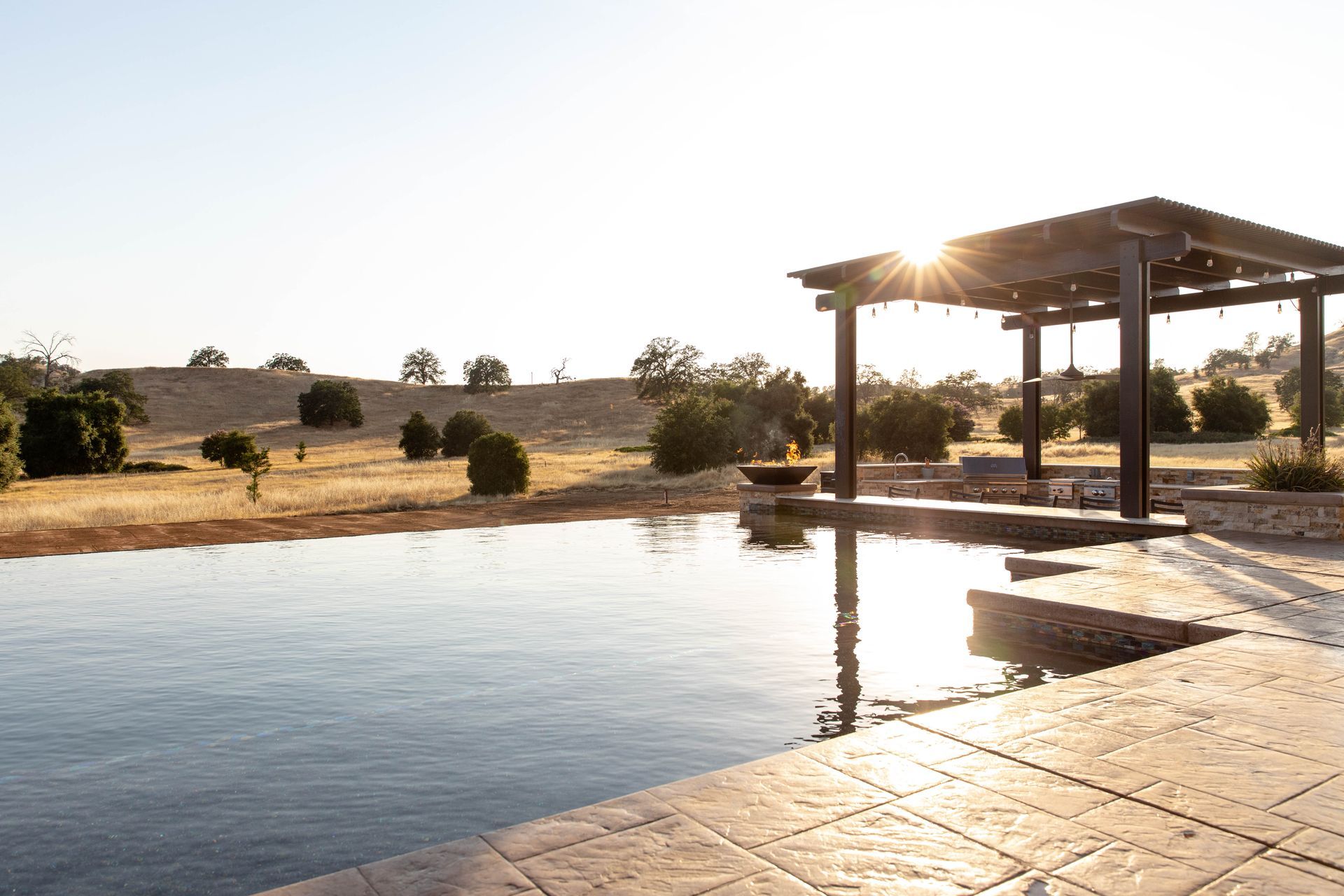 Sunlit resort pool beside a shaded patio overlooking dry grassland and scattered trees