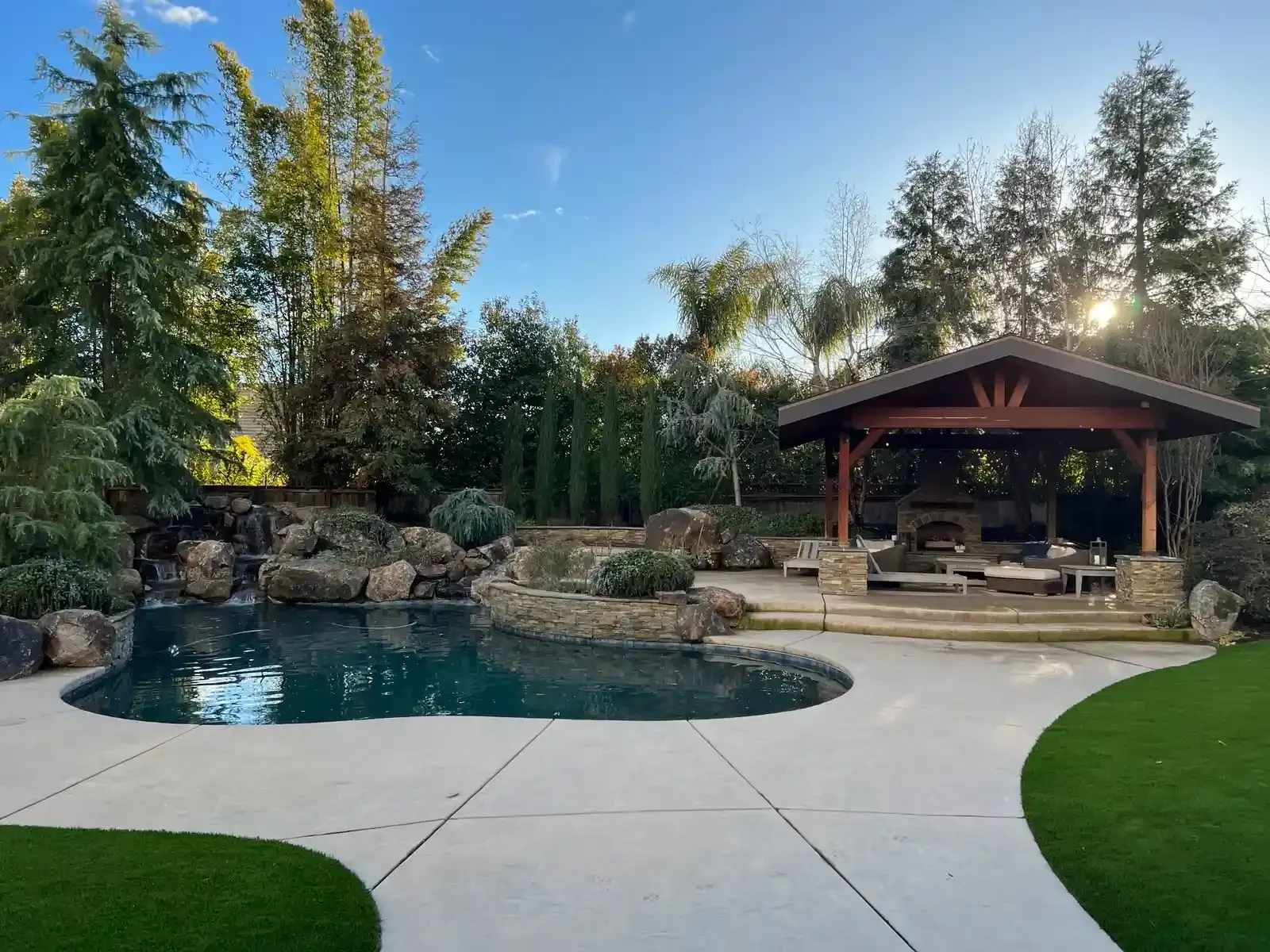 Backyard pool with stone waterfall, patio, and covered seating area surrounded by trees
