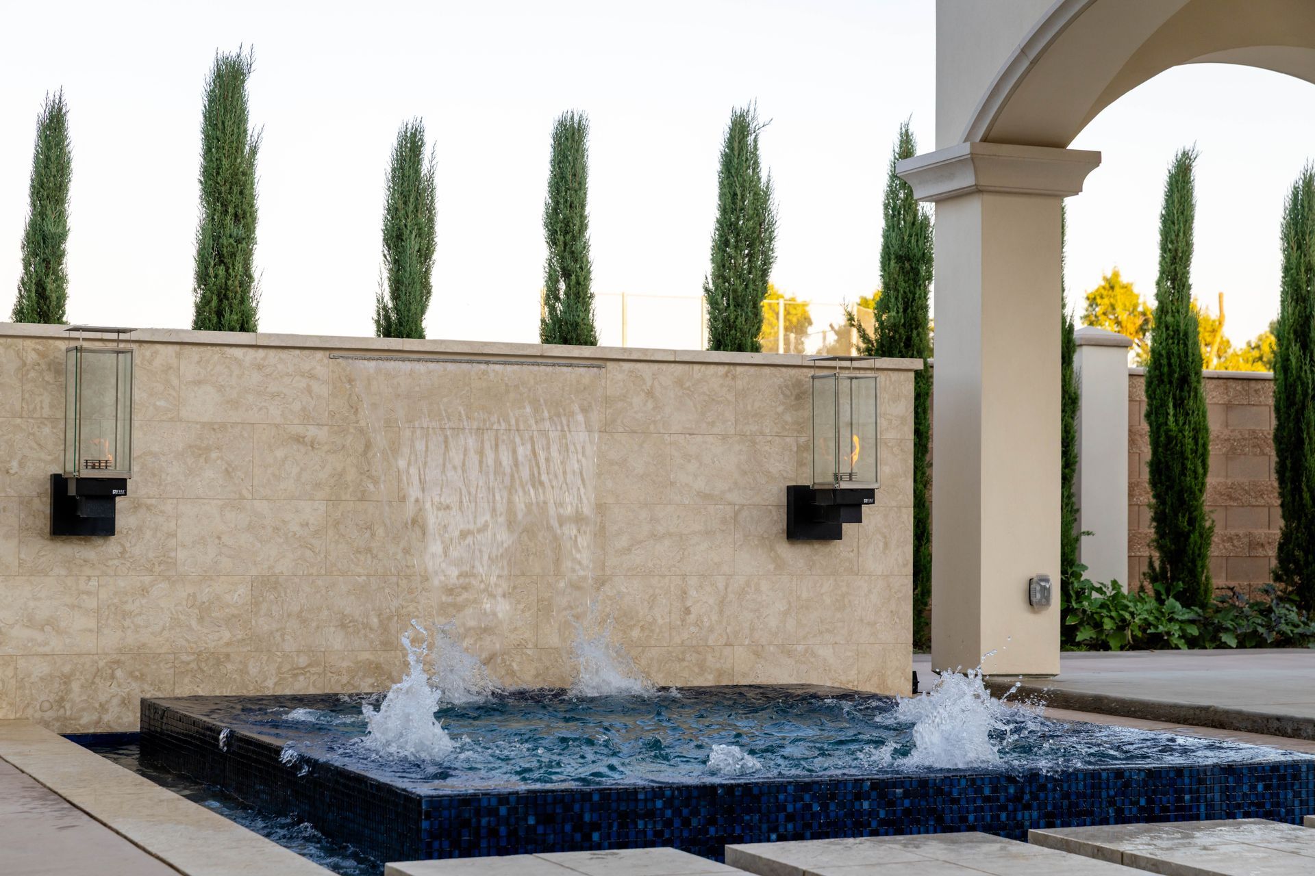 Courtyard fountain with water jets, beige stone wall, and tall cypress trees behind