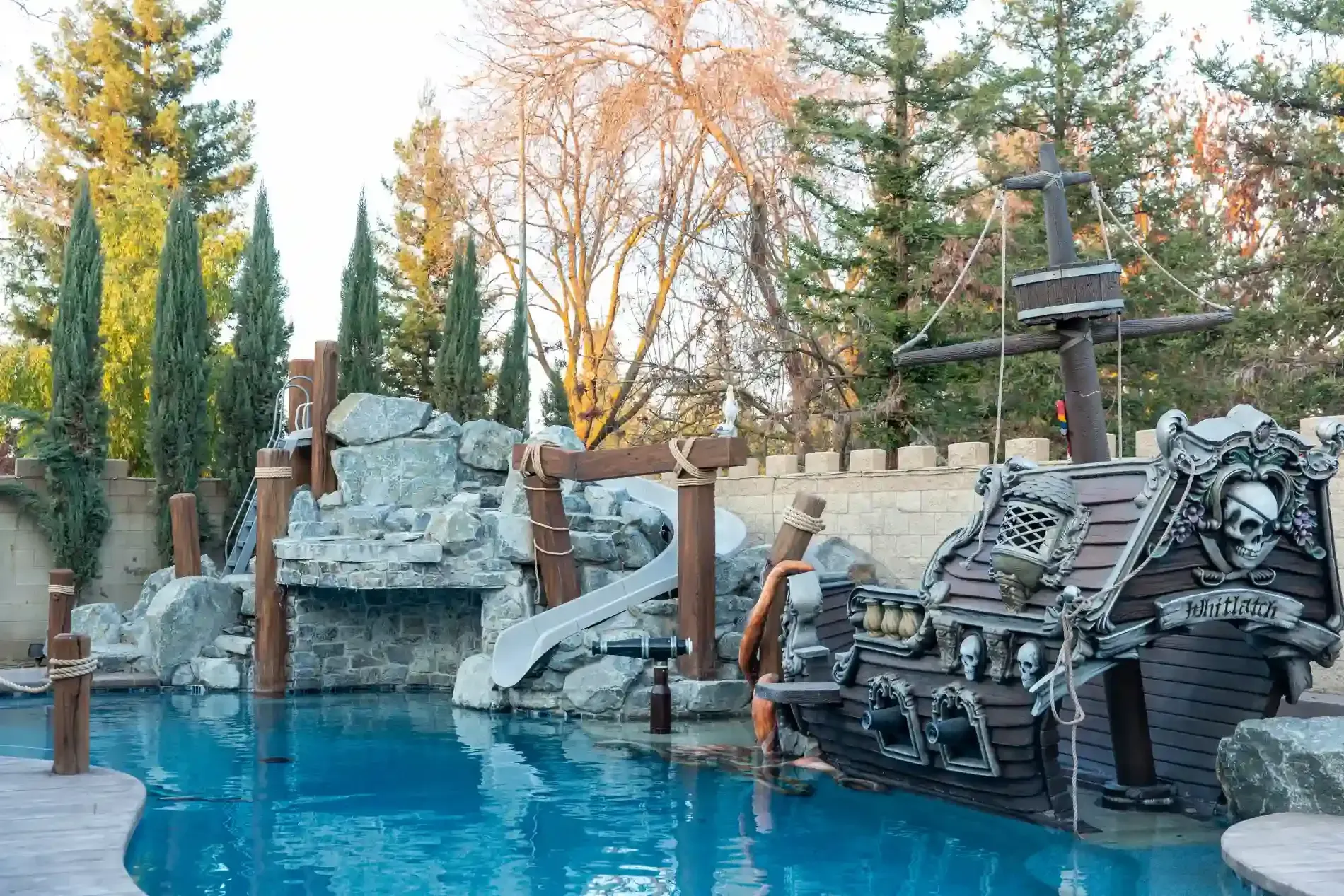 Rocky poolside water slide with blue water, stacked lounge chairs, and tall trees in the background