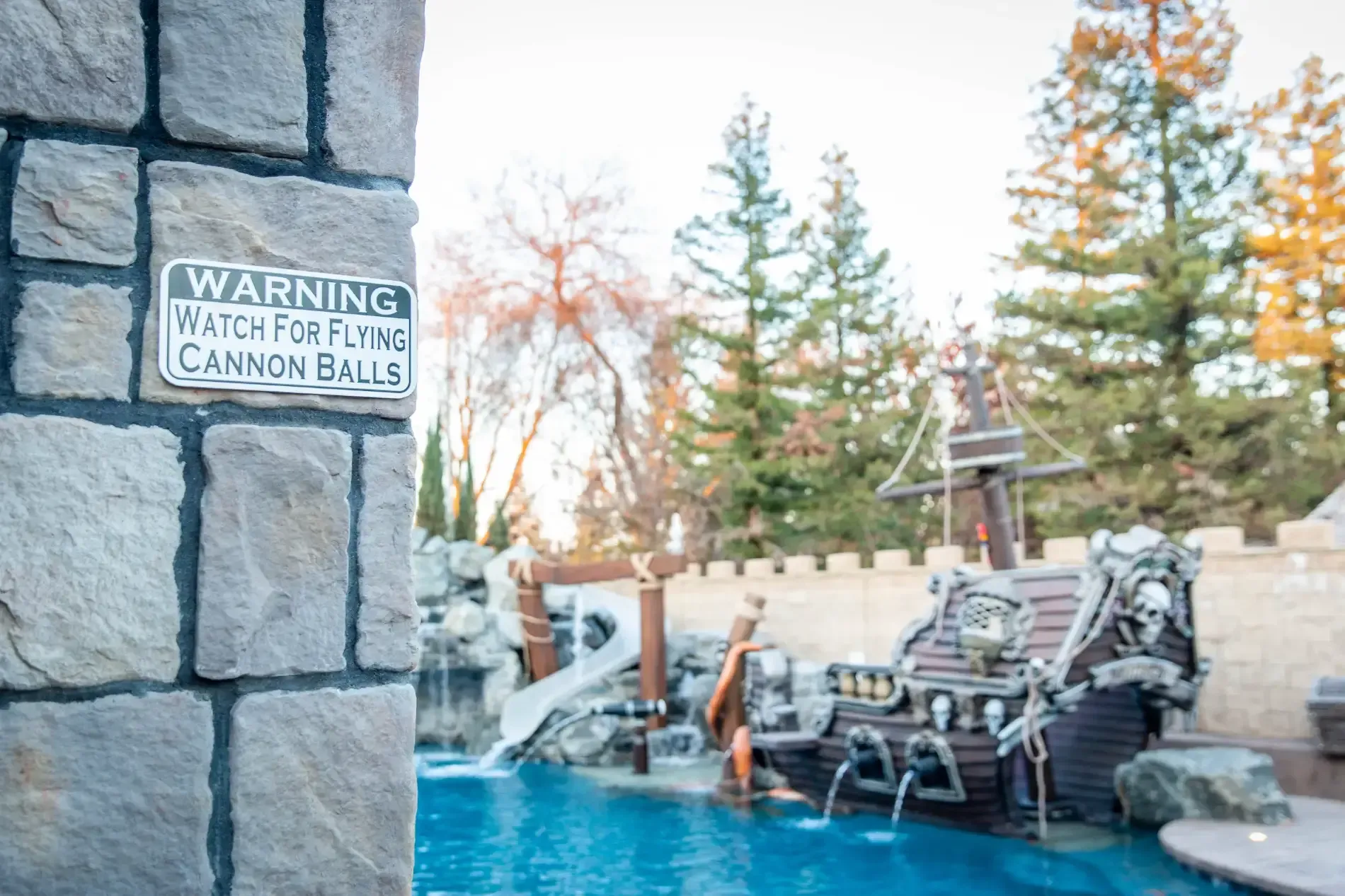 Pool with rock wall and cannonball warning sign, lined with chairs and tall trees.