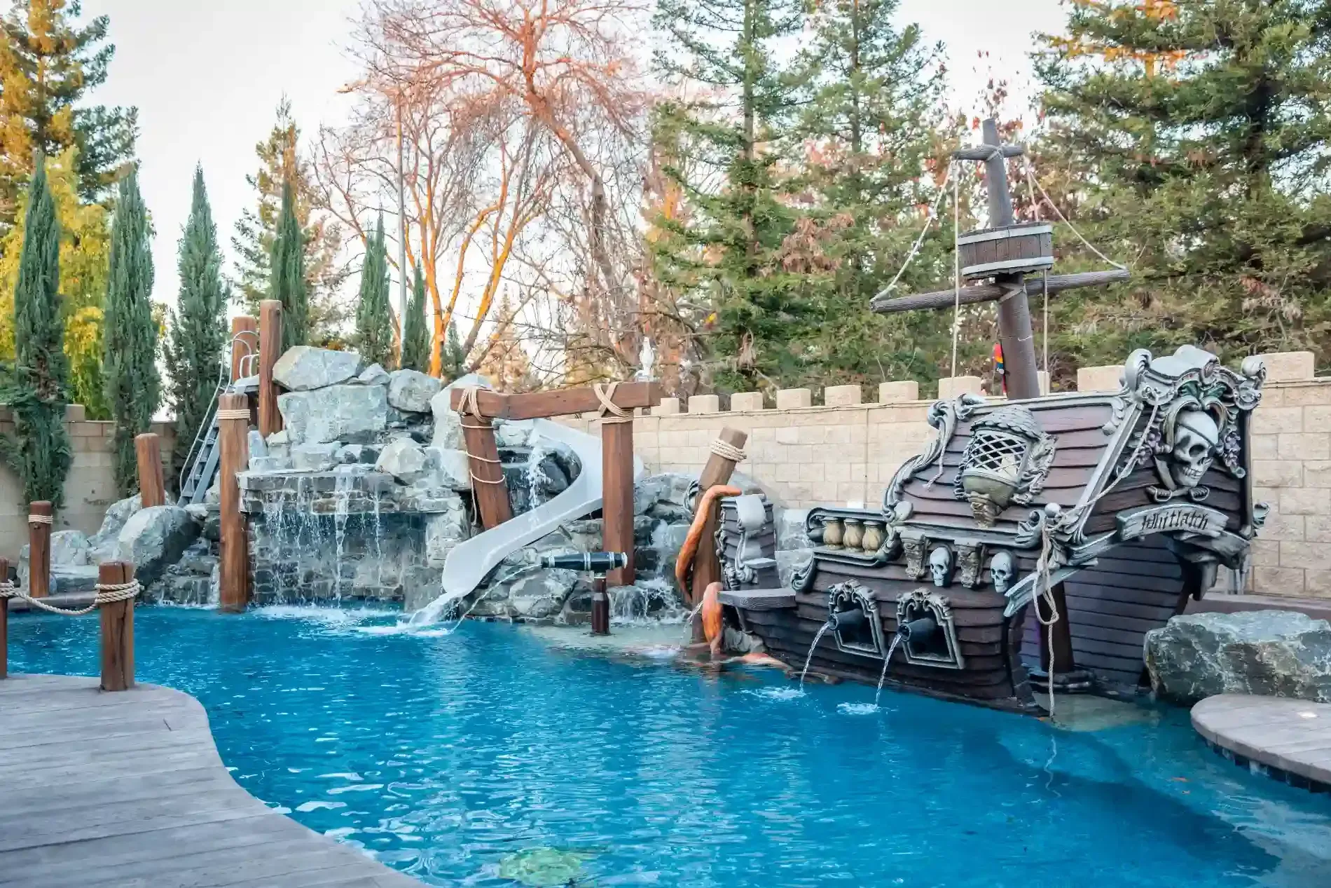 Blue pool with pirate ship play structure, rock waterfall, and autumn trees in the background