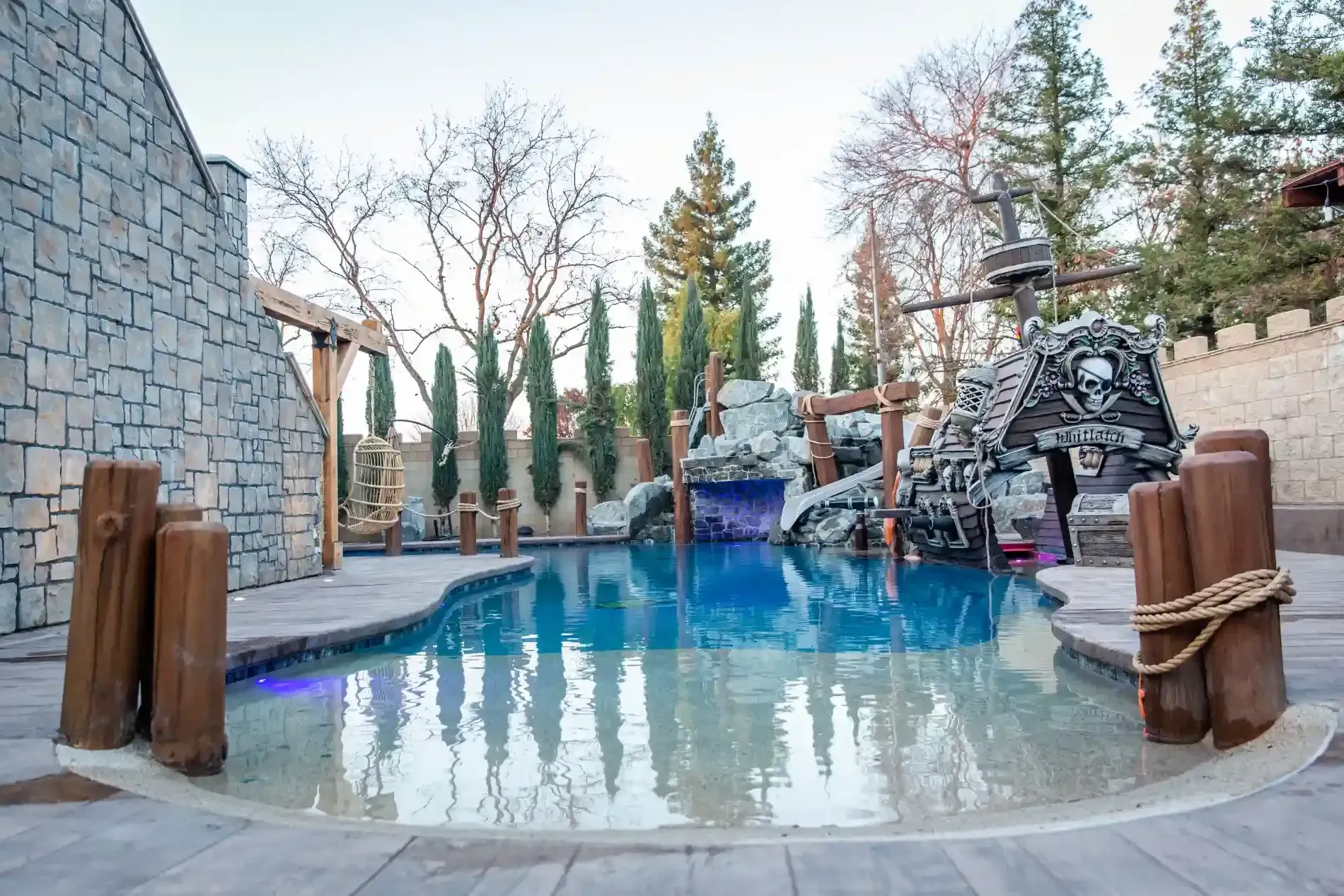 Stone courtyard pool with blue water, wooden posts, and a small rock waterfall surrounded by tall trees
