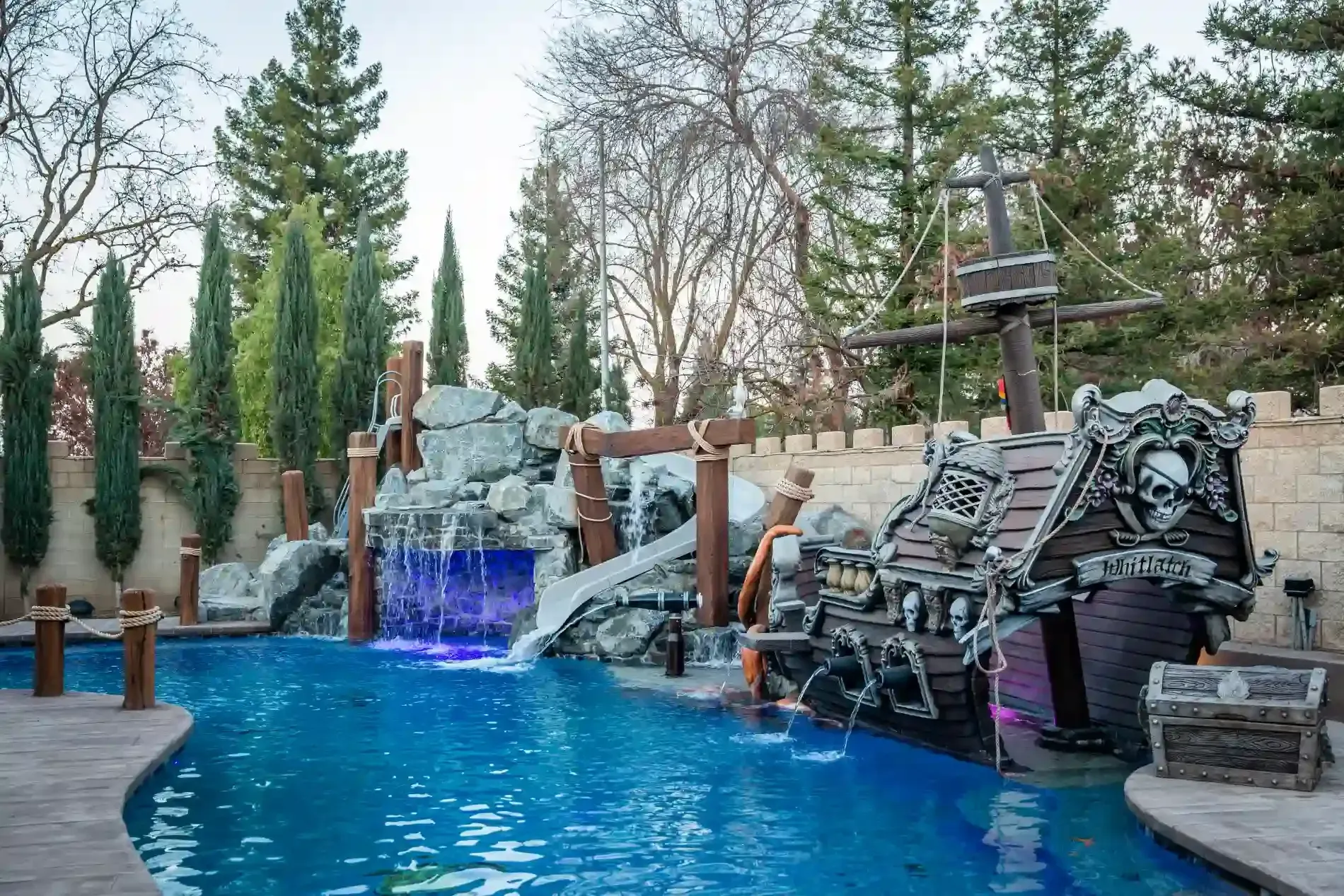 Backyard pool with blue water, rock waterfall, and stacked lounge chairs among tall trees