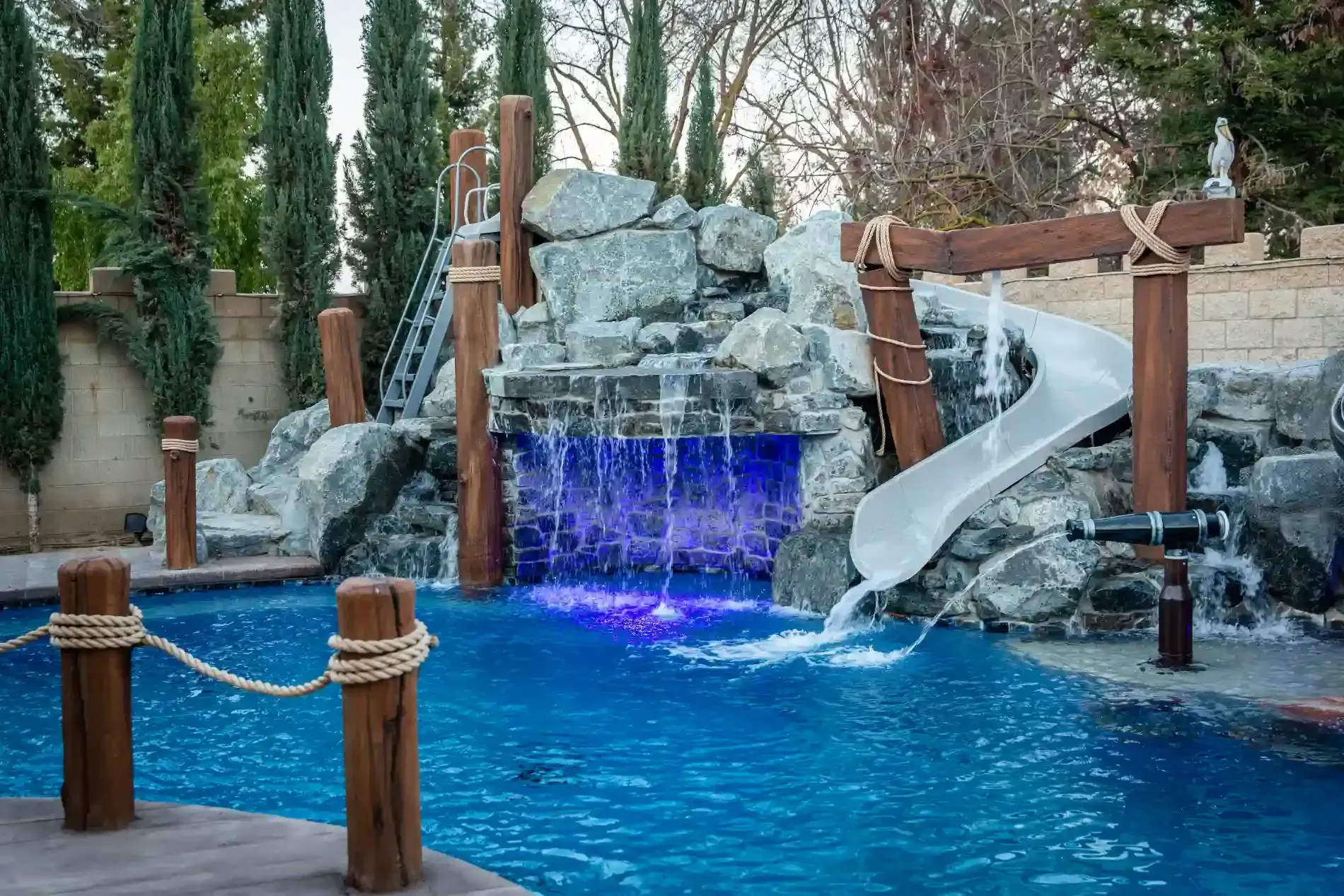 Rock waterfall and blue pool with a lit purple cascade and a water slide in a resort setting