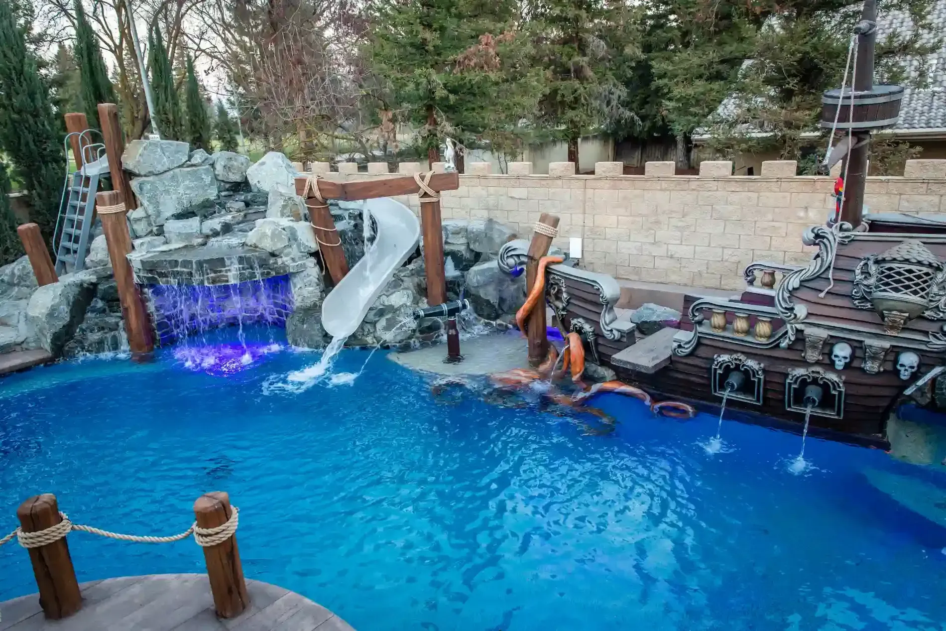 Swimming pool with rock waterfall, blue lighting, and lounge seating beside a stone wall.