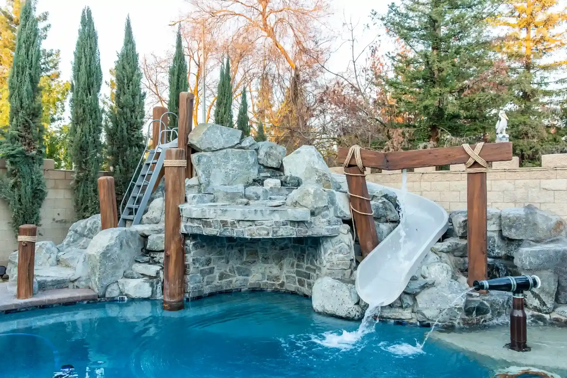 Rock waterfall into a blue pool with a slide, surrounded by trees and stone landscaping