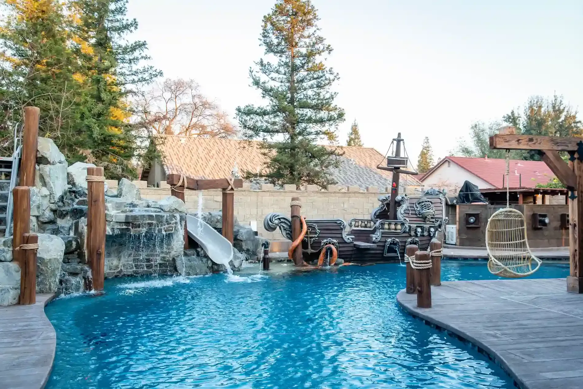 Resort pool with waterfalls, stone deck, and lounge chairs surrounded by trees and buildings