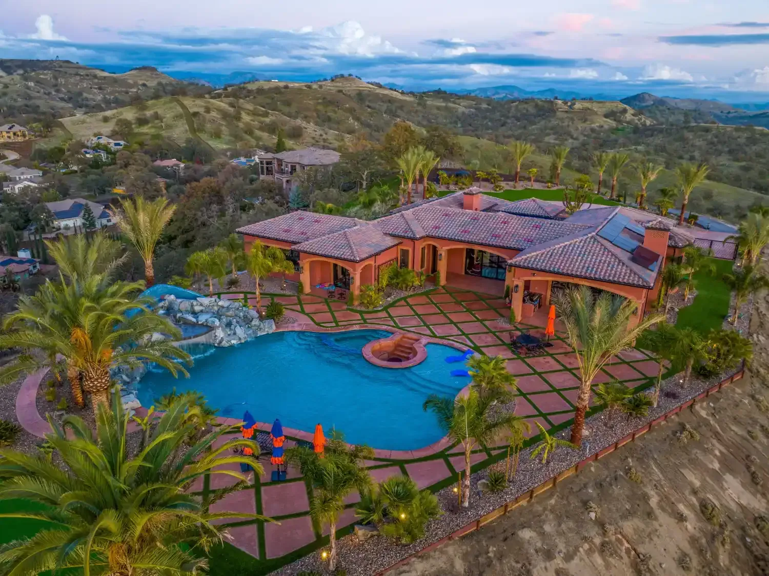 Aerial view of a villa with a blue pool, palm trees, and mountain landscape beyond.