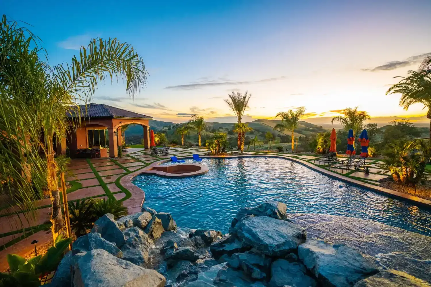 Luxury backyard pool at sunset with rock waterfall, palm trees, and patio seating