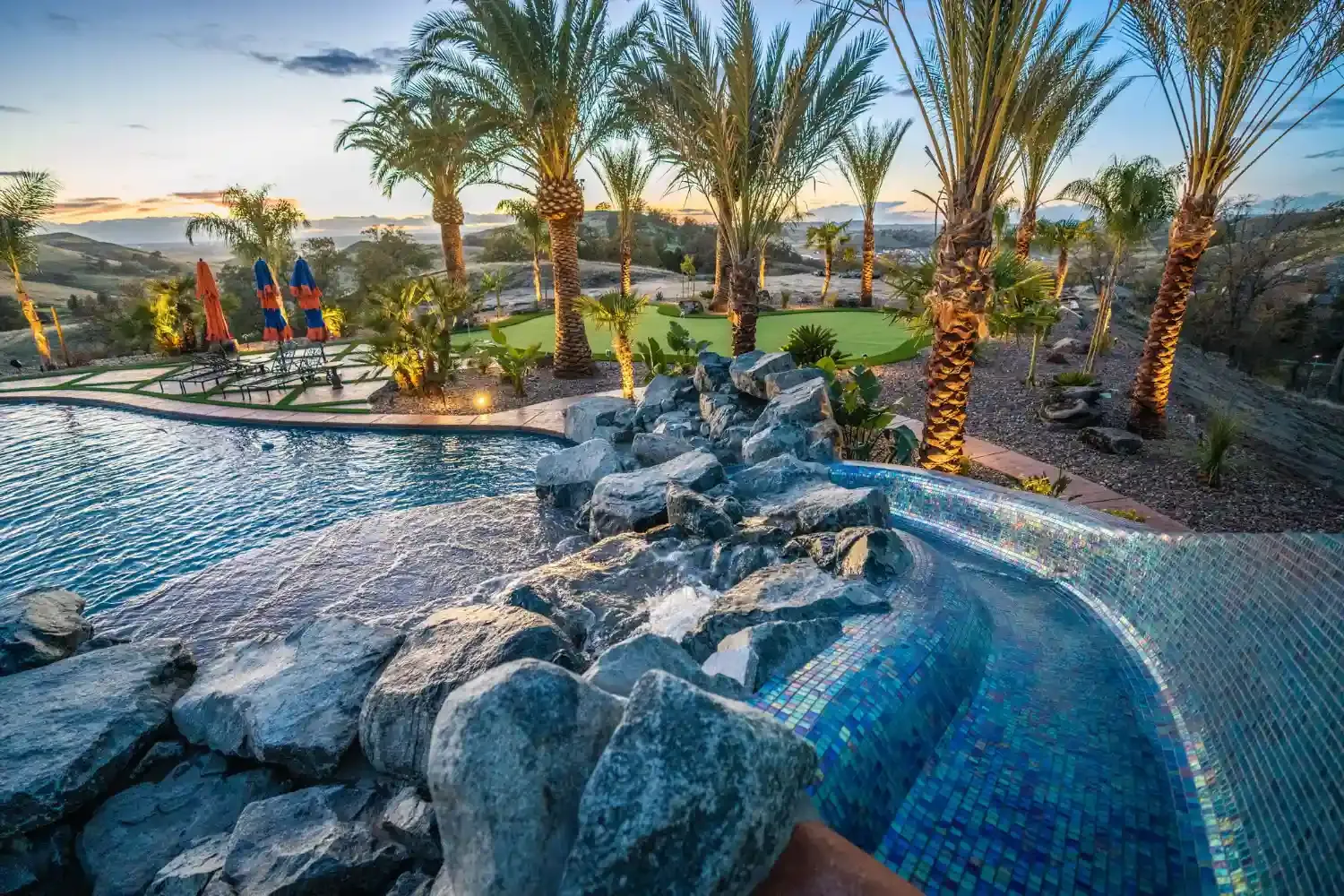 Resort pool with rock waterfall, palm trees, lounge chairs, and colorful umbrellas at sunset