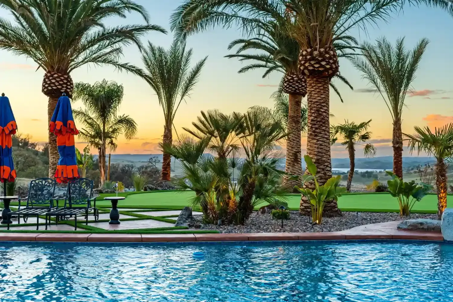 Resort pool with palm trees, lounge chairs, and colorful umbrellas at sunset
