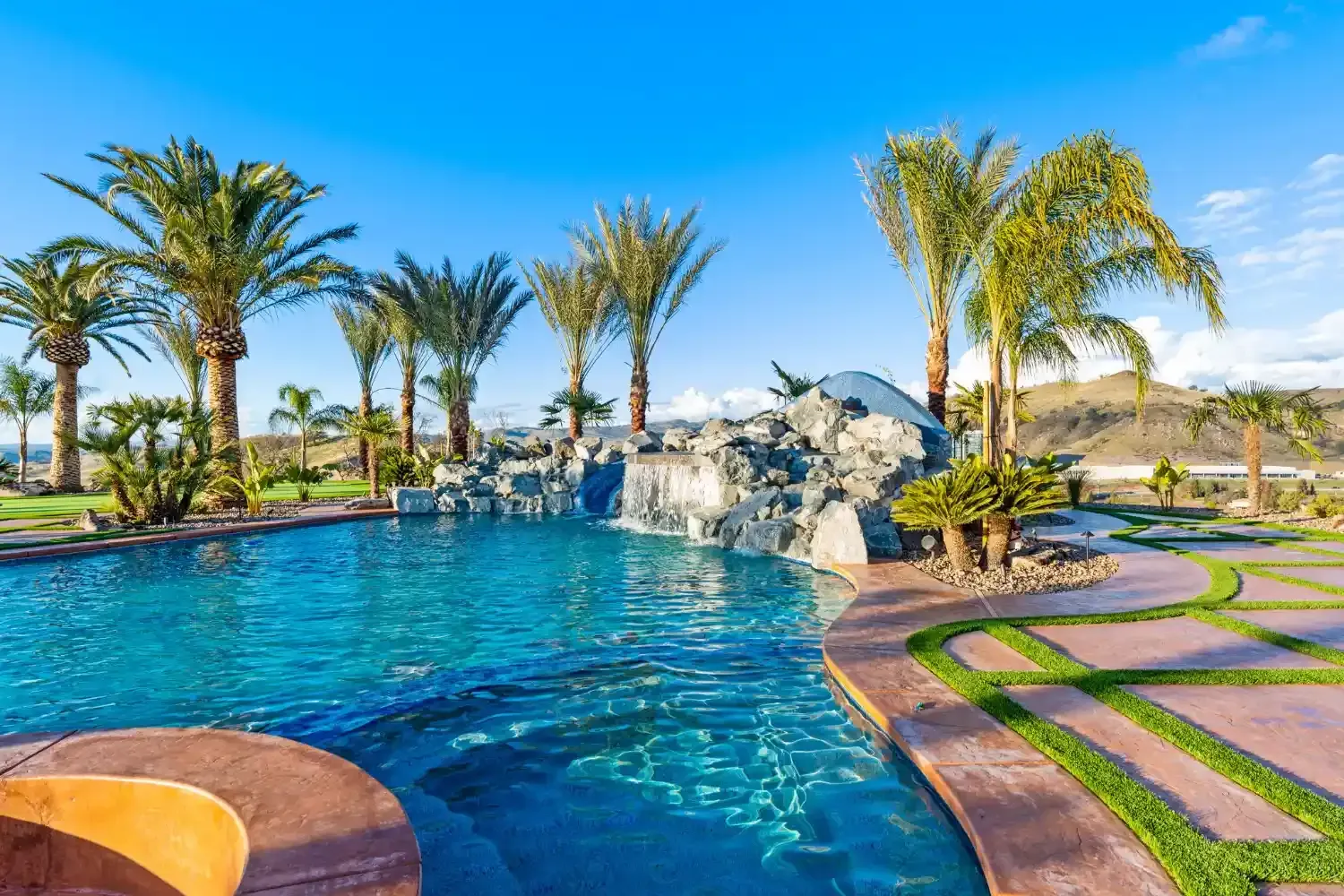 Resort-style pool with palm trees, rock waterfall, and curved stone path under a clear blue sky