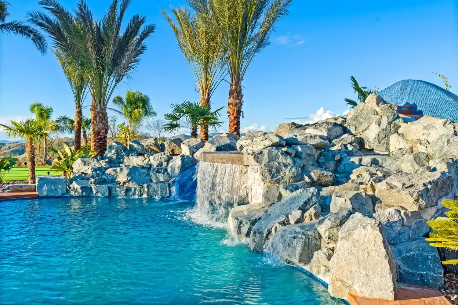 Resort pool with turquoise water, palm trees, and a rocky waterfall under a clear blue sky