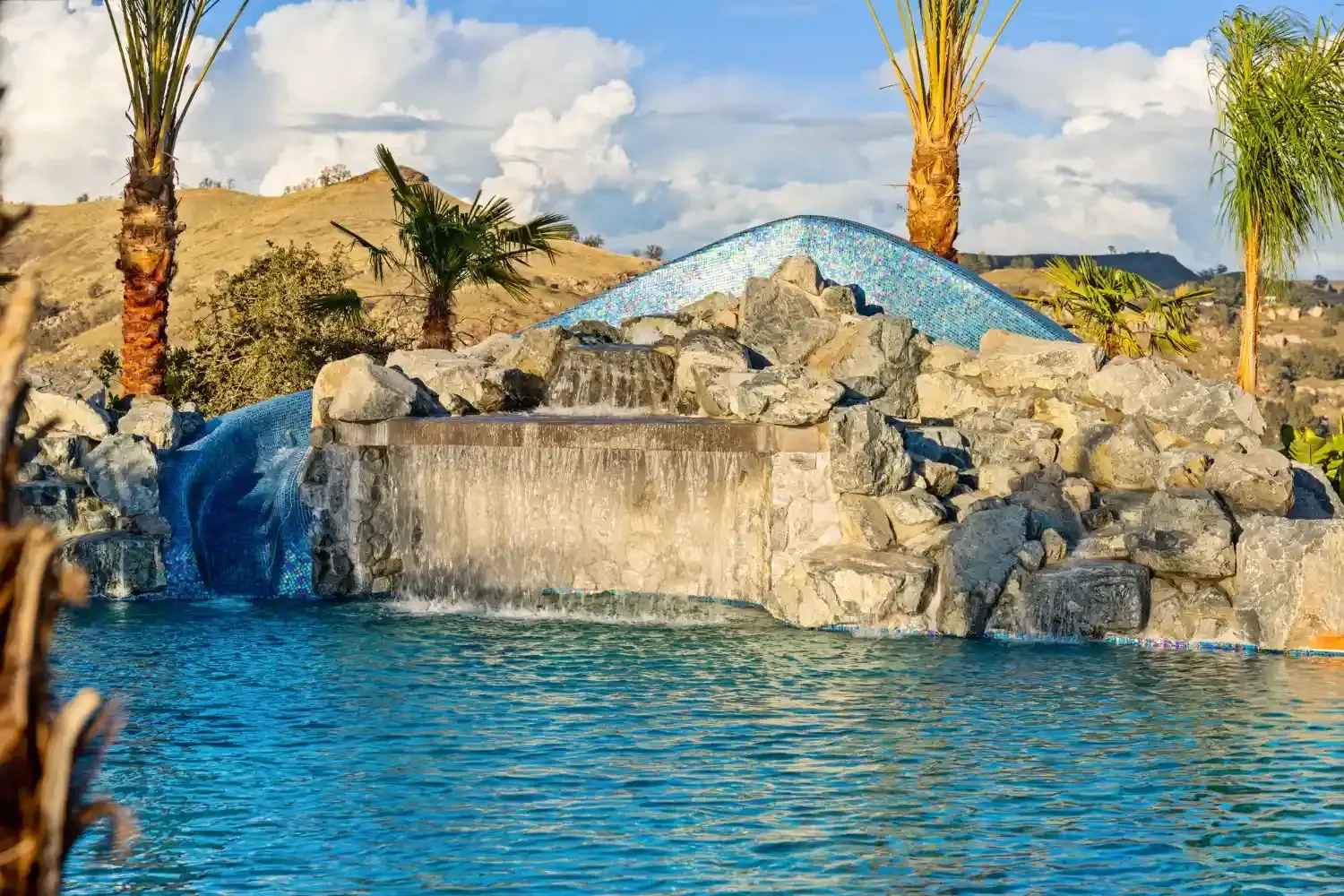 Rock waterfall pool with blue water, rocks, and palm trees under a sunny sky