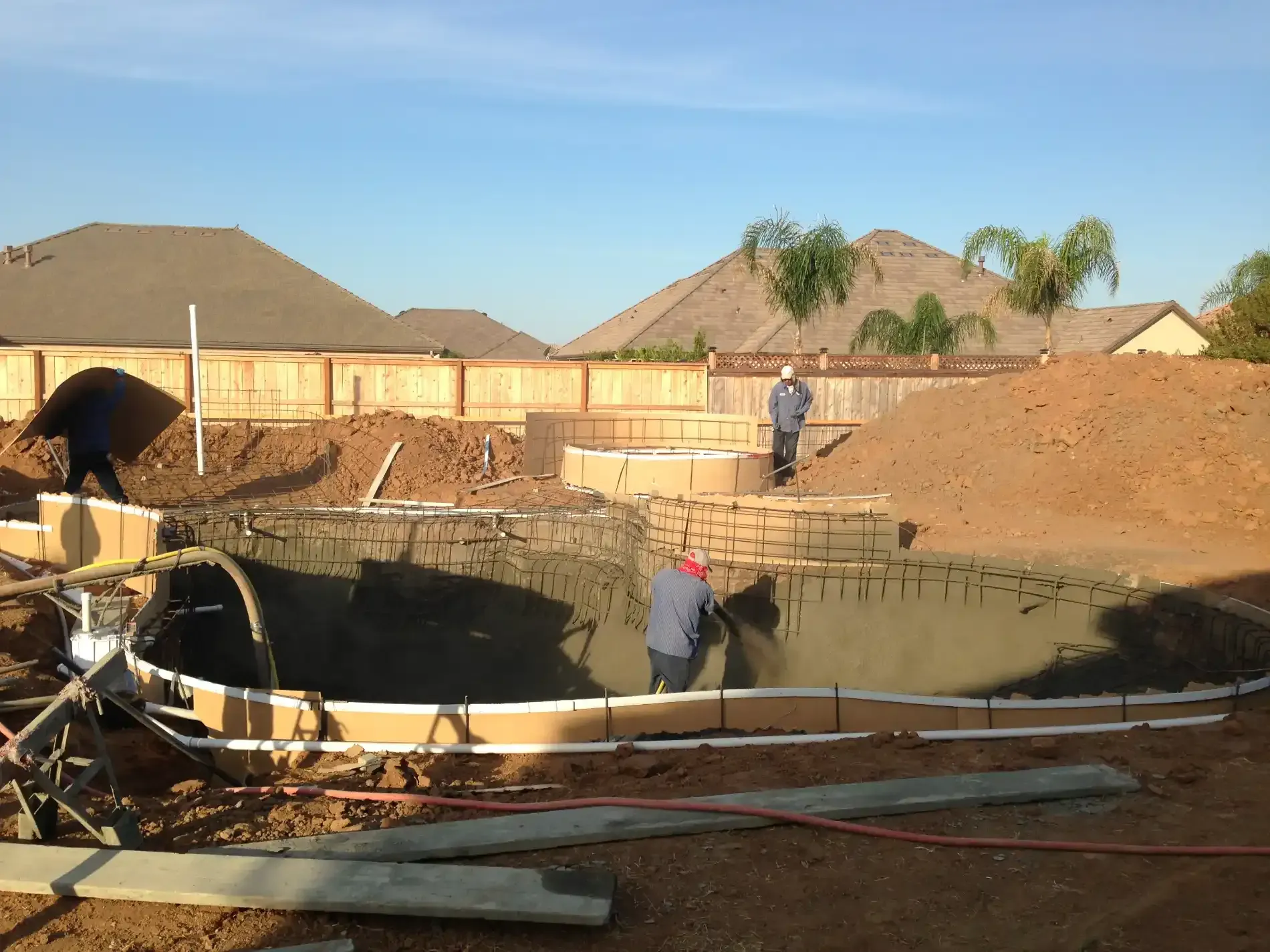 Construction site with a large in-ground pool excavation and workers near piles of dirt and wood framing