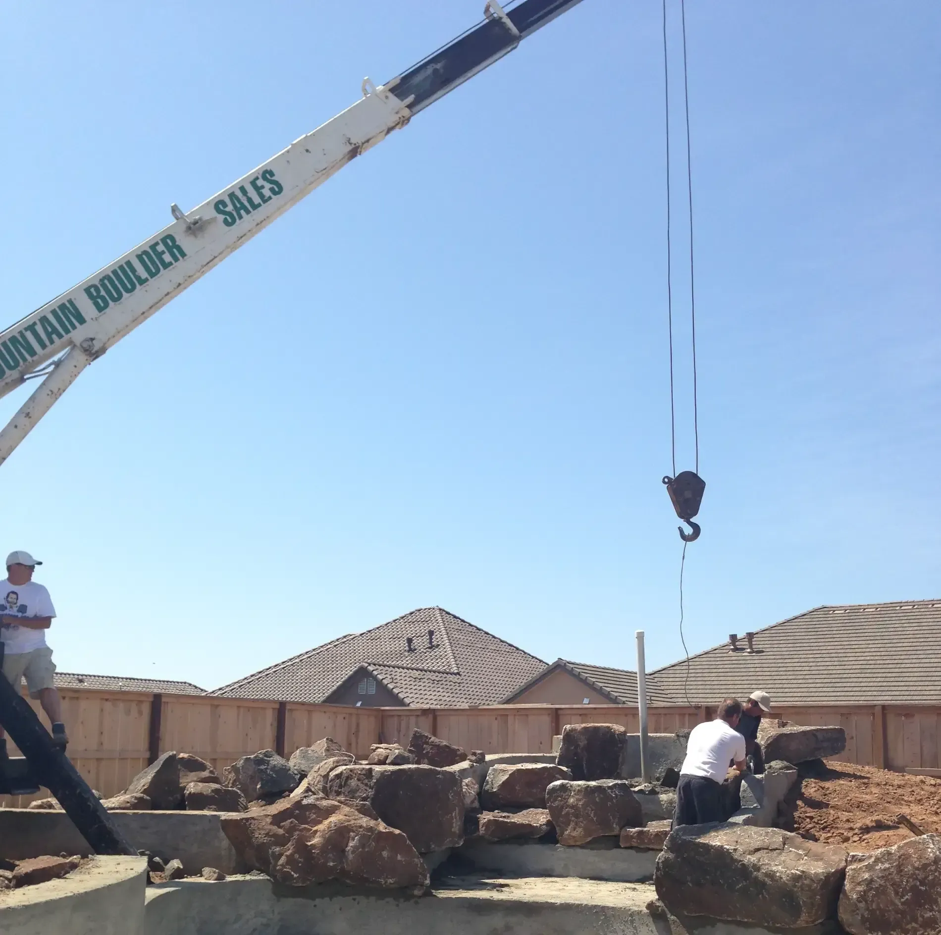 Crane lifting a large boulder at a rocky construction site with workers nearby and houses in the background