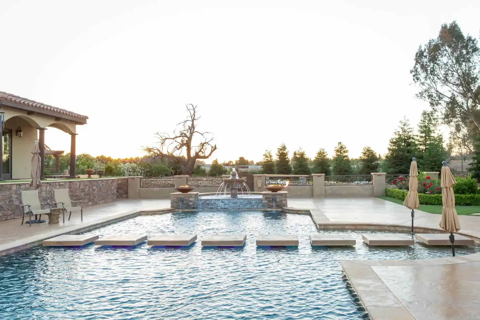 Modern resort pool with a waterfall, stone patio, and sunset view beyond the water