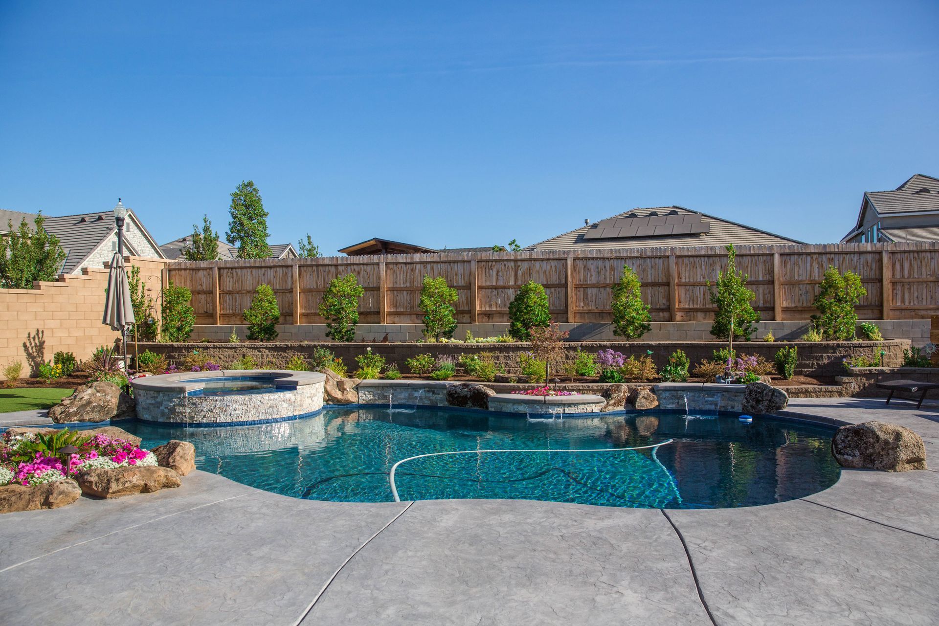 Backyard with a curved blue swimming pool, stone spa, and landscaped garden under a clear sky