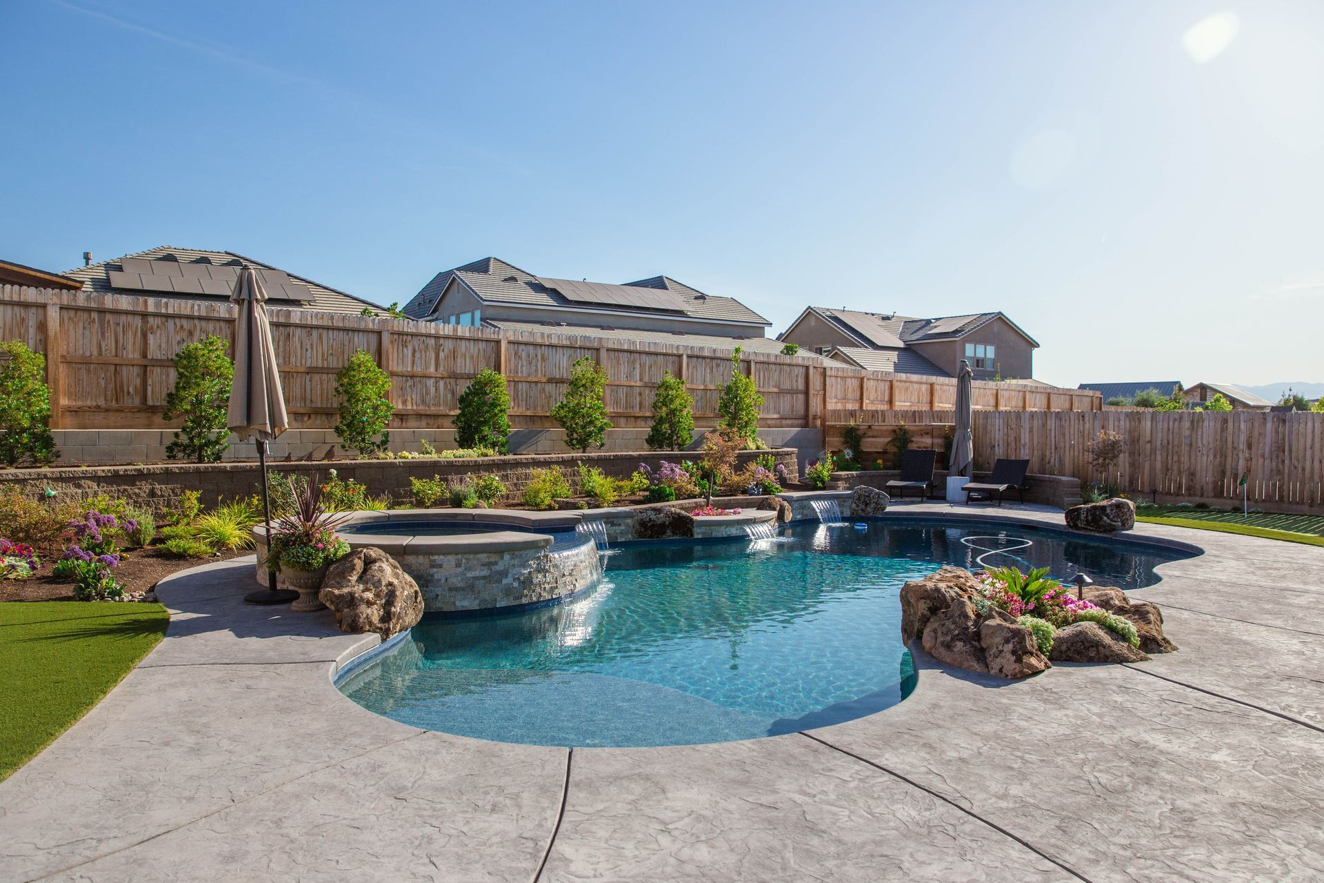 Backyard swimming pool with rock waterfall, concrete patio, and wooden privacy fence under clear sky