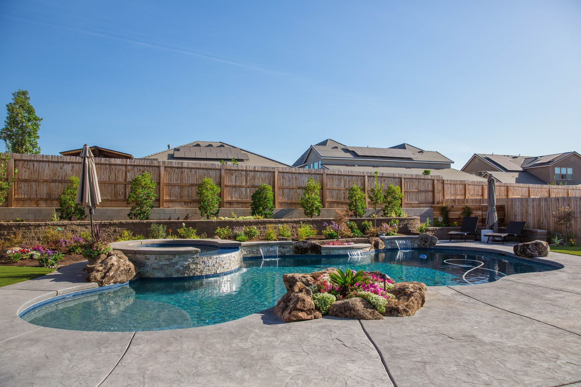 Backyard swimming pool with spa, rock landscaping, and wooden fence under a clear blue sky