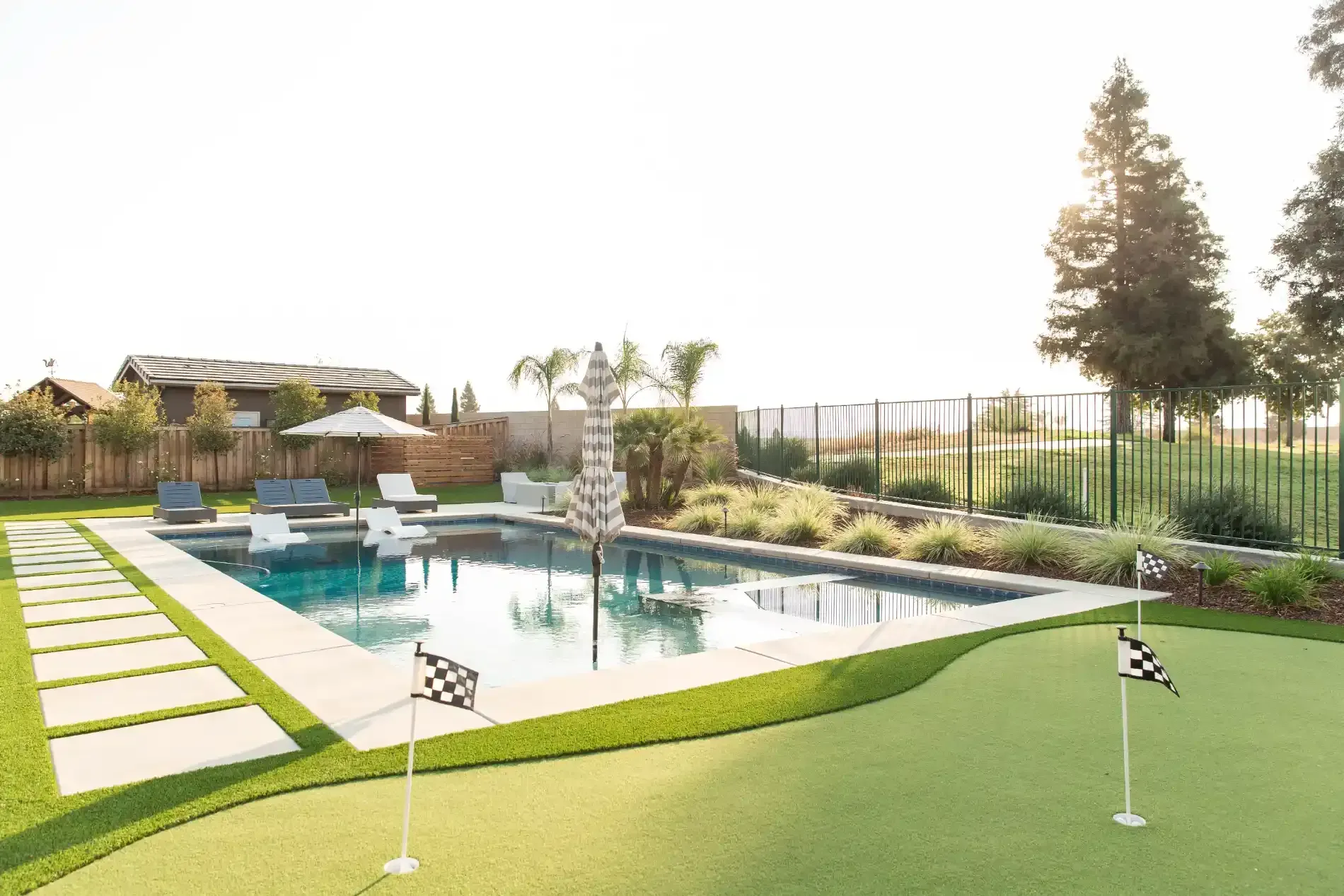 Modern backyard pool with green lawn, stepping stones, patio seating, and trees at sunset