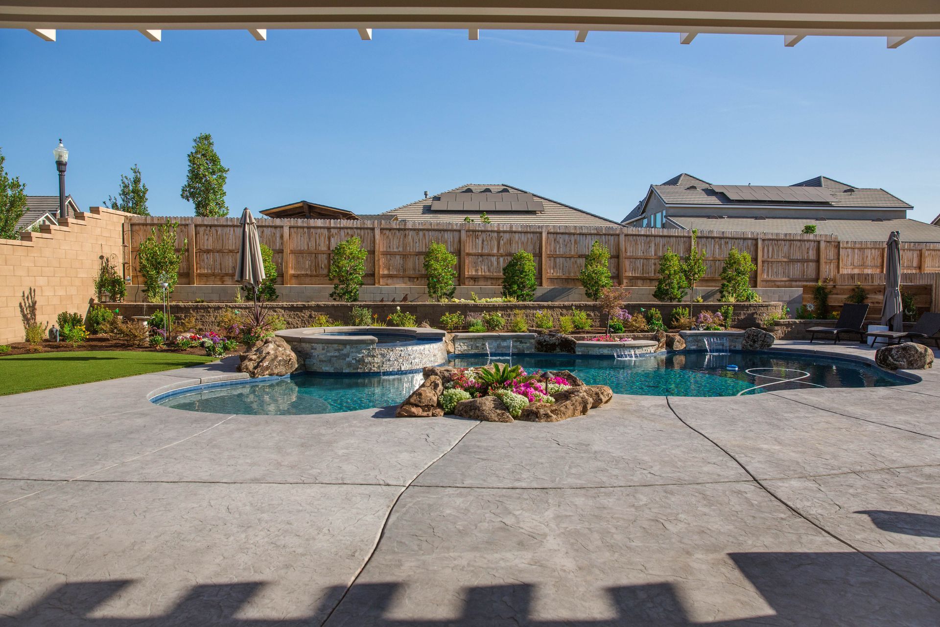 Backyard with a swimming pool, stone waterfall, flower bed, and paved patio under a blue sky