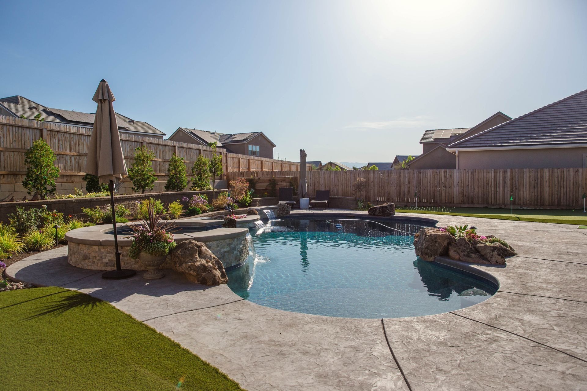 Backyard pool with stone patio, landscaping, and sunlit suburban houses in the background