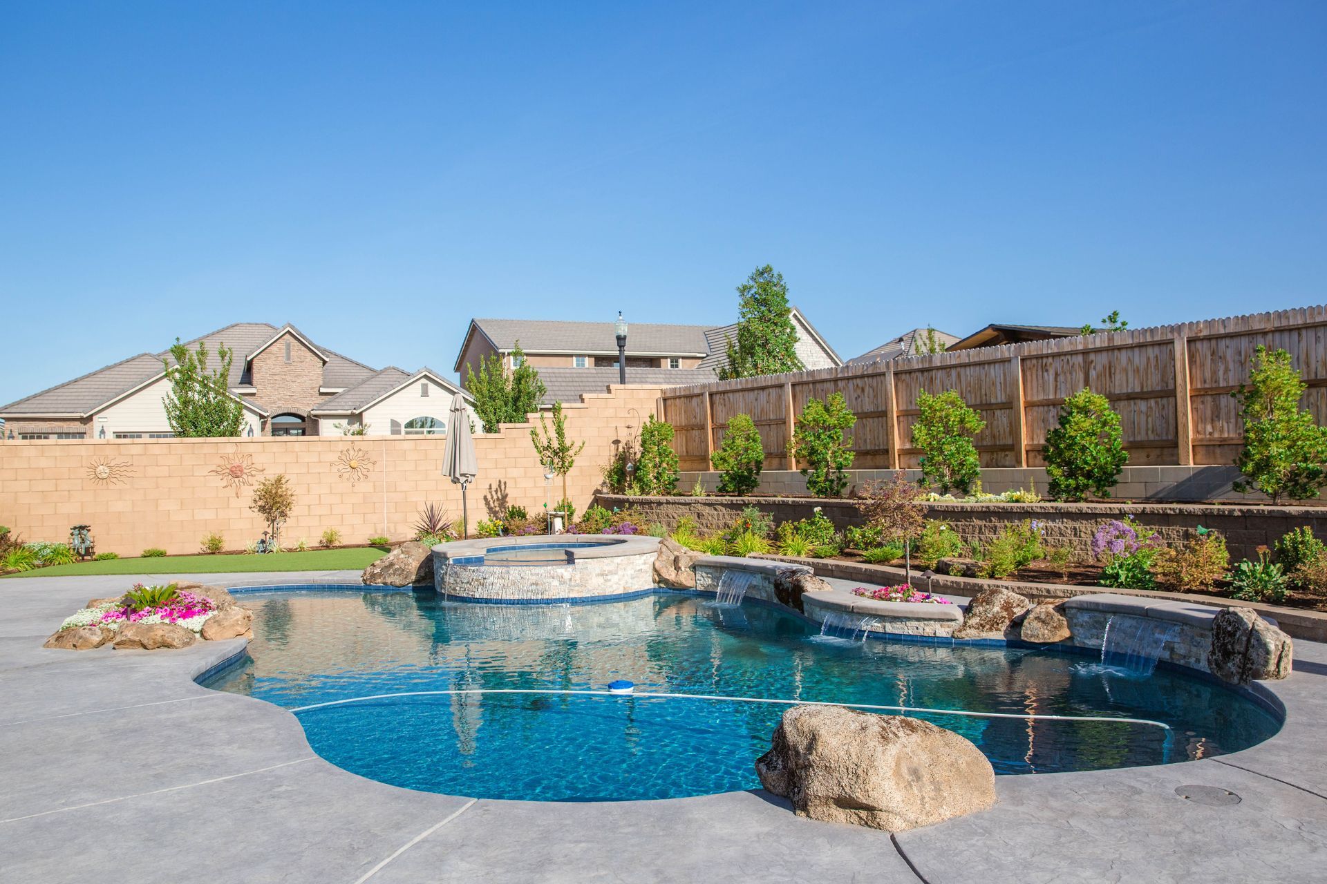 Backyard swimming pool with rock waterfall, patio, and fenced landscaping under a clear blue sky