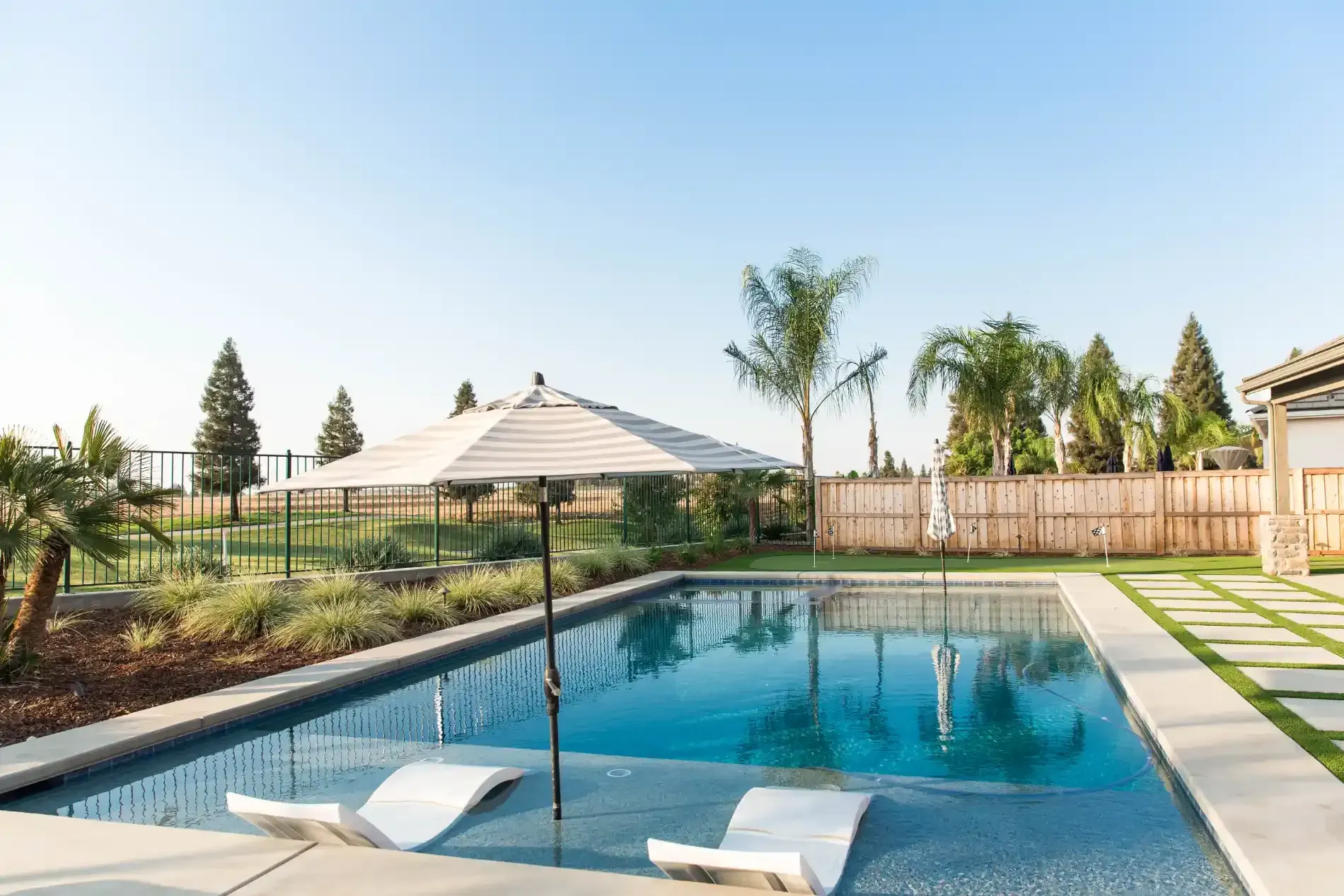 Backyard swimming pool with lounge chairs, patio umbrella, and palm trees under a clear sky