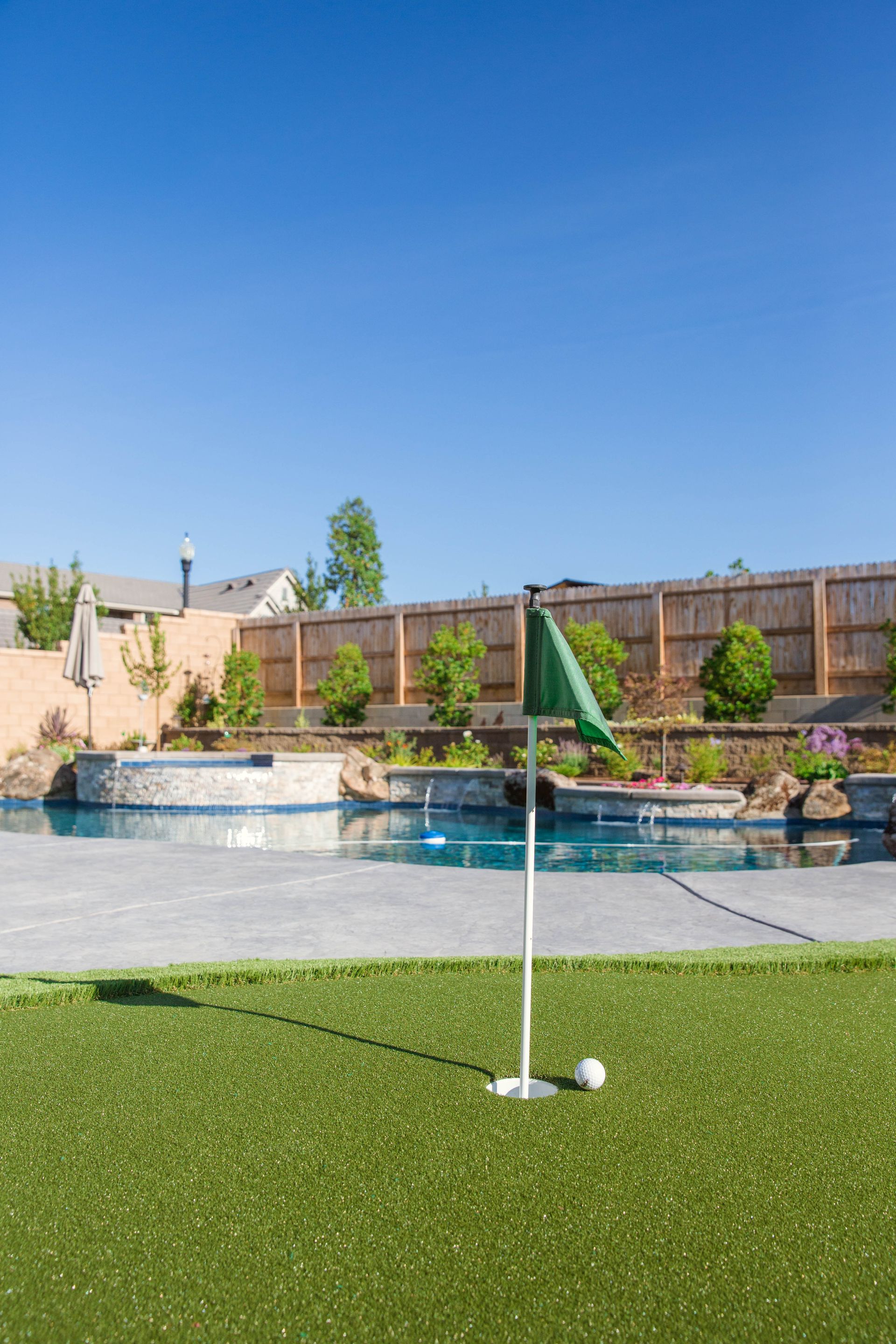 Golf ball and flag on artificial turf beside a backyard pool under a clear blue sky