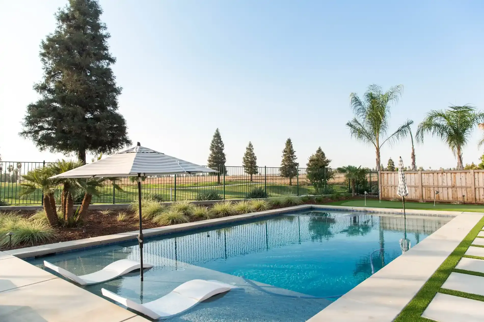 Modern backyard pool with white lounge chair, umbrella, and landscaped garden under a clear sky