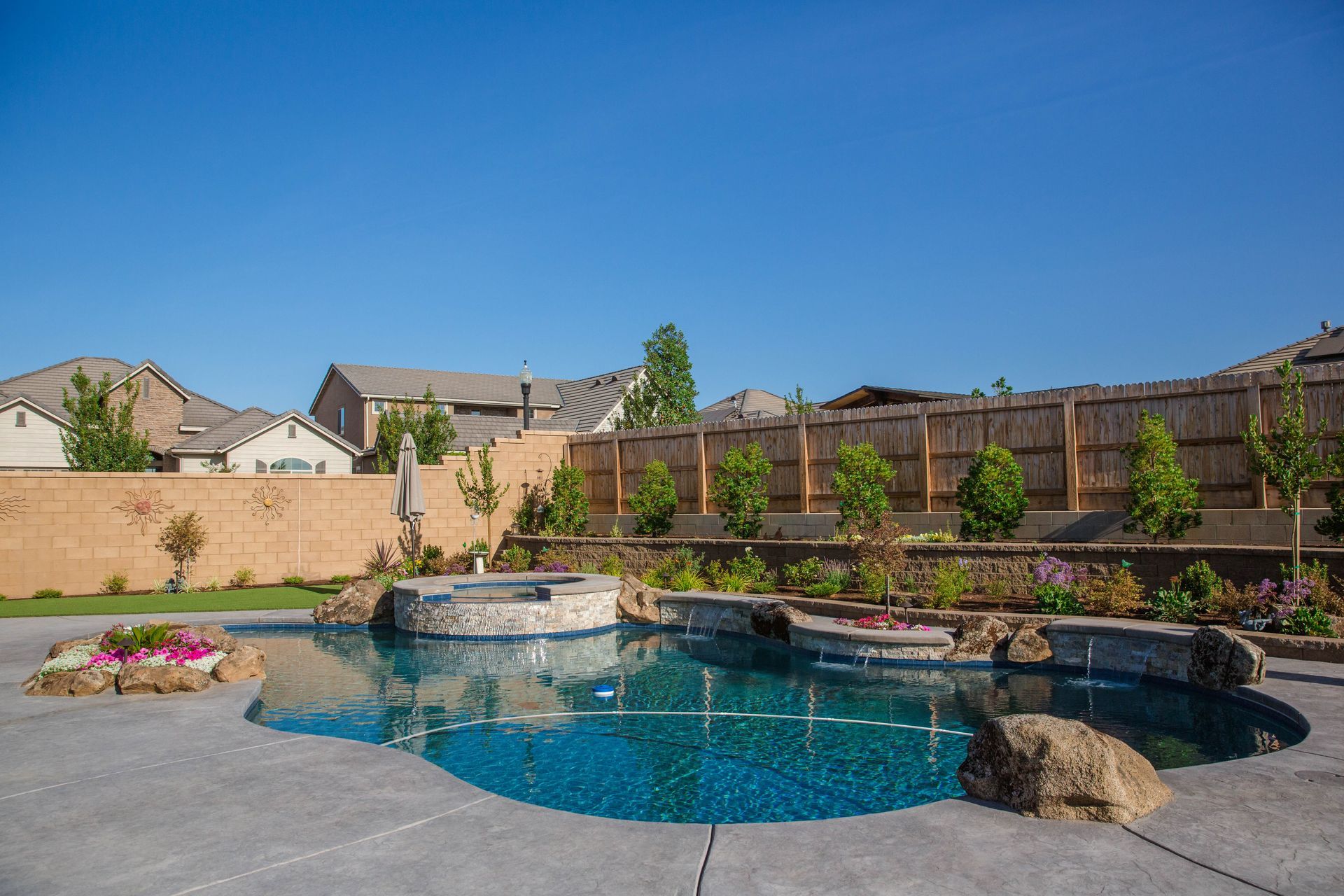 Backyard pool with stone waterfall, blue water, patio, and wooden fence under a clear sky