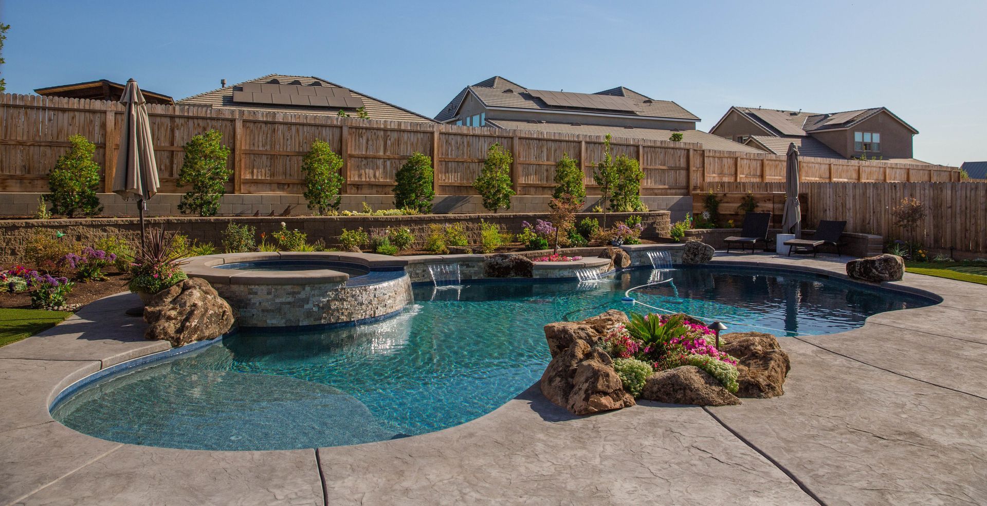 Backyard pool with rock waterfall, blue water, patio, and desert-style landscaping under a sunny sky