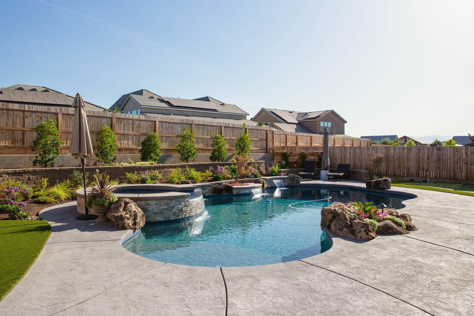 Backyard swimming pool with rock waterfall, patio, and fenced suburban homes in the background