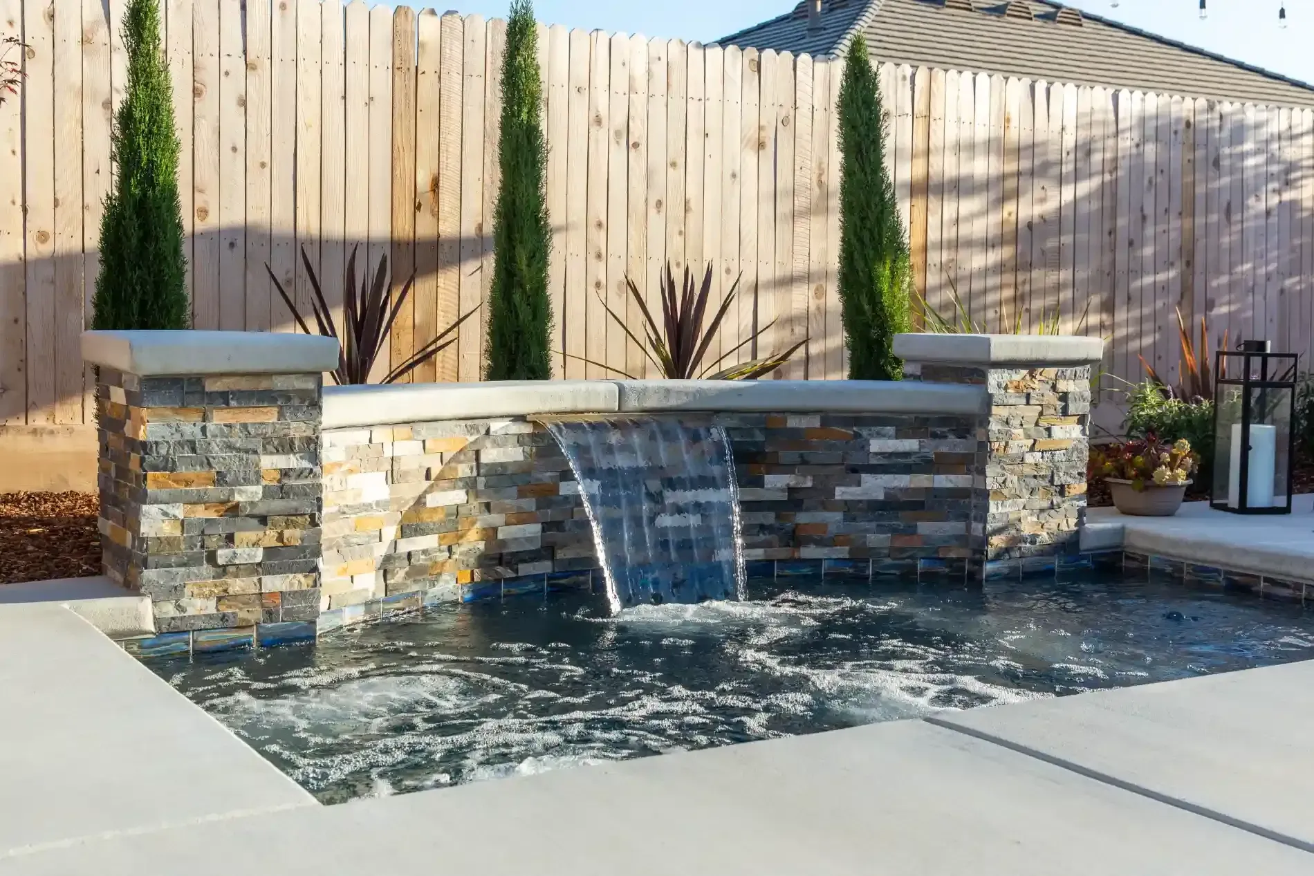 Modern backyard stone water feature with cascading waterfall and tall evergreens on a patio