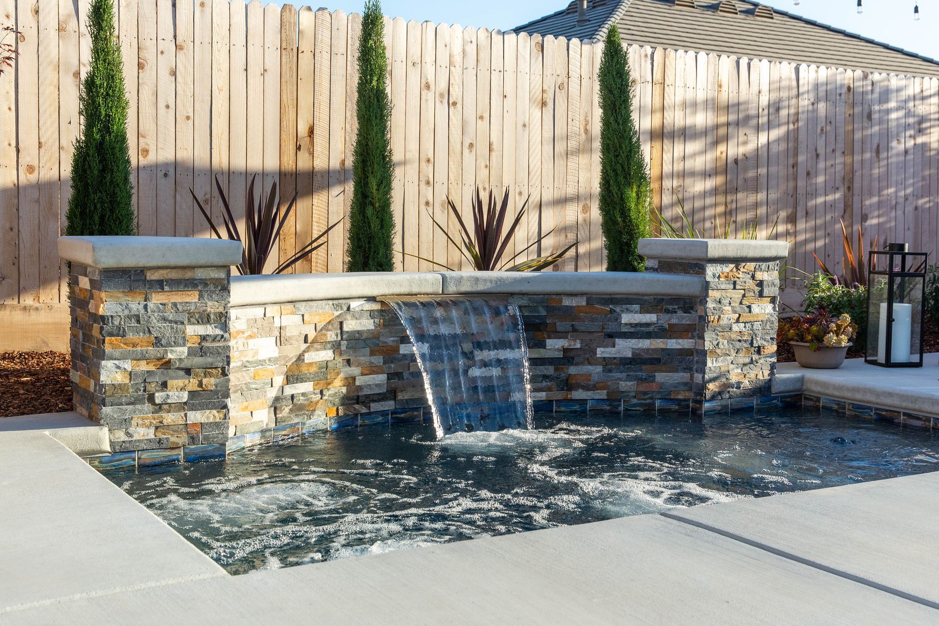 Backyard stone water feature with waterfall into a shallow pool, bordered by tall cypress trees.