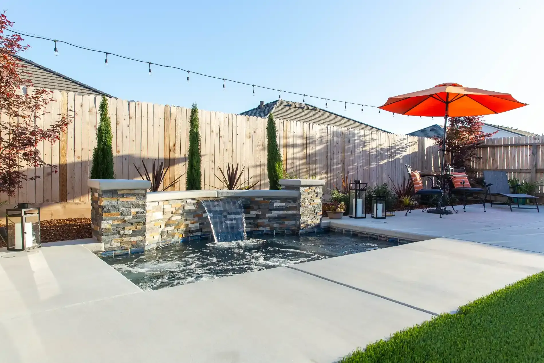 Backyard patio with stone fire pit, string lights, orange umbrella, and lounge chairs under a clear sky