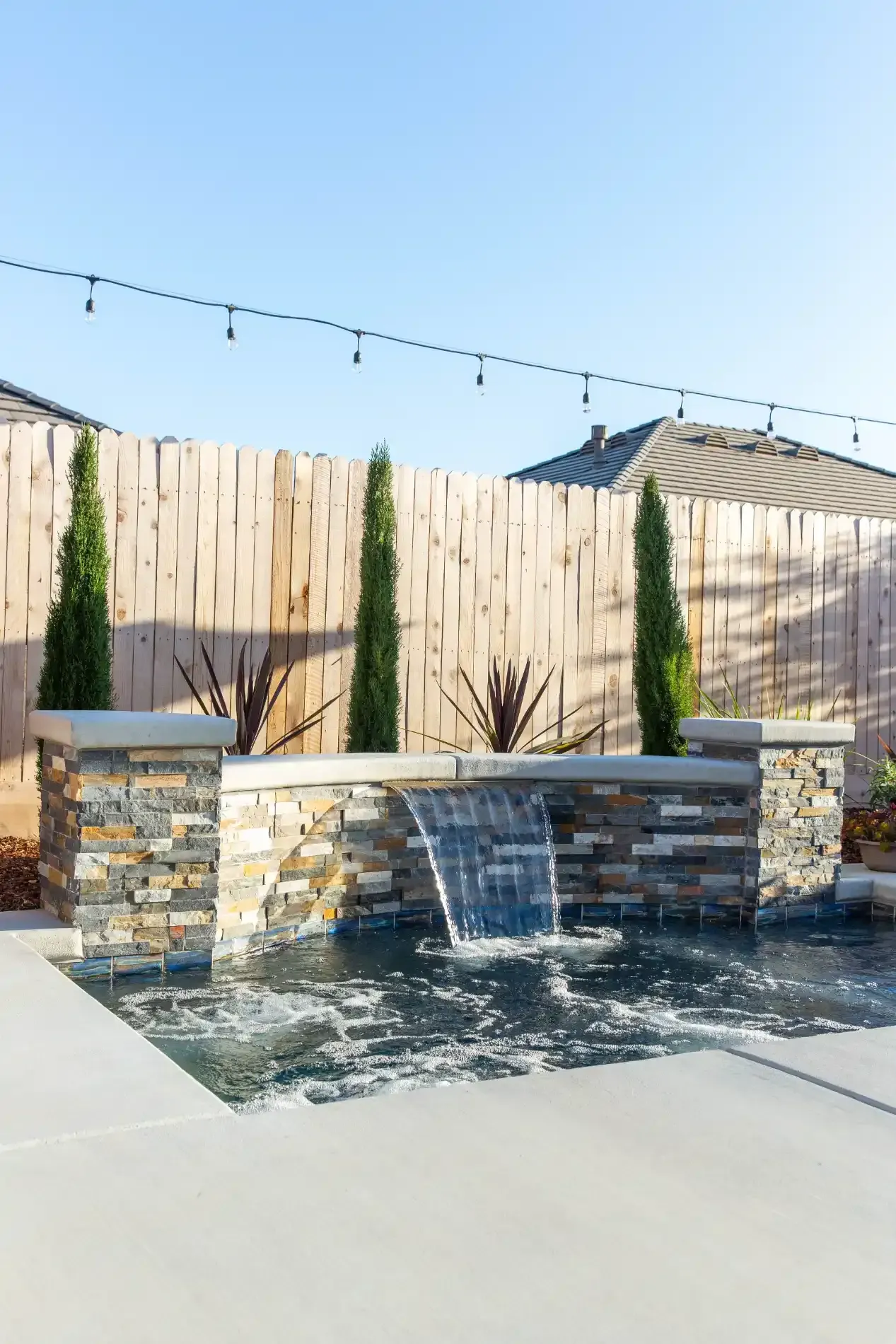 Backyard fountain with water spilling over stone ledges beside a wooden fence and string lights