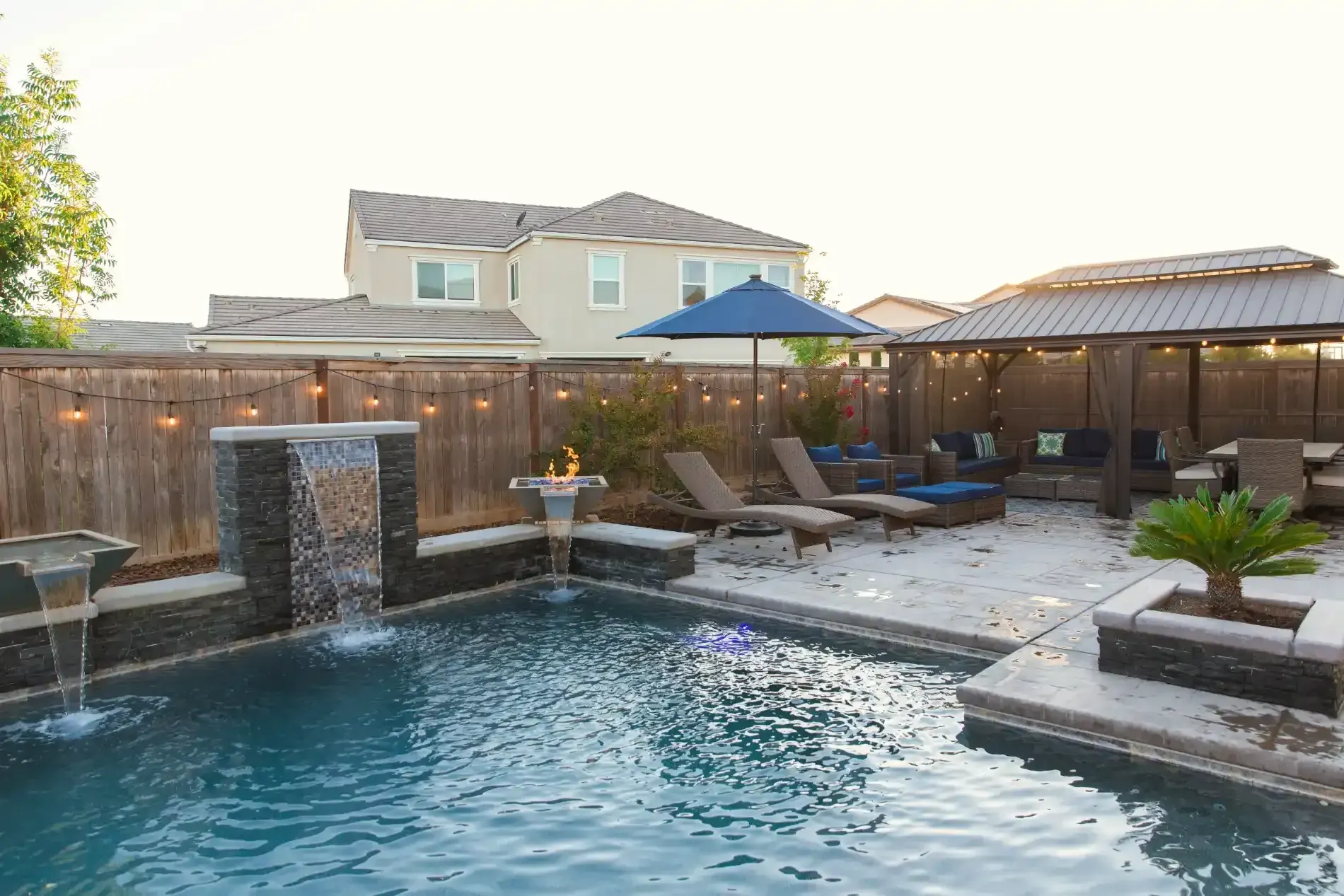 Backyard pool with waterfall, lounge chairs, umbrella, and modern house at dusk