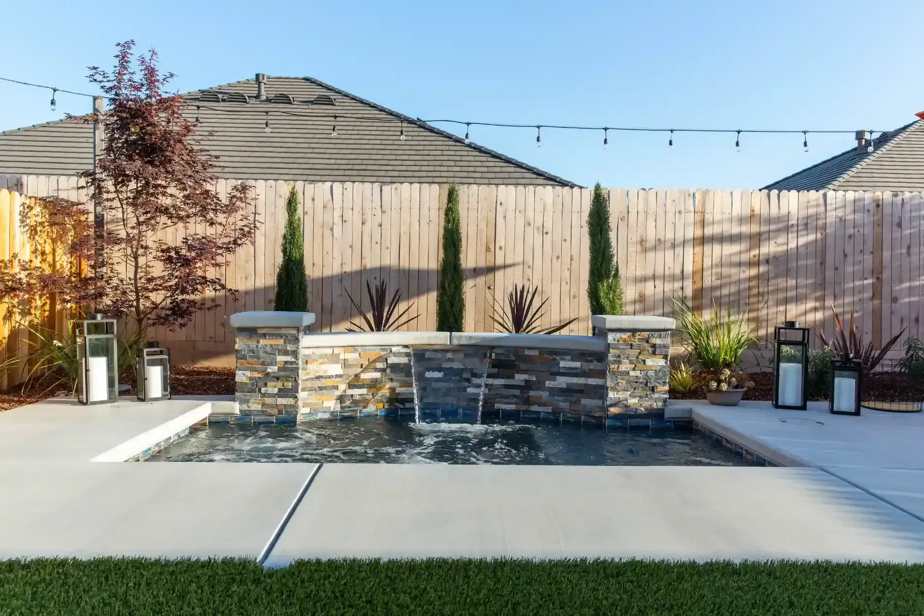 Backyard patio with stone fire pit, string lights, potted plants, and wooden privacy fence
