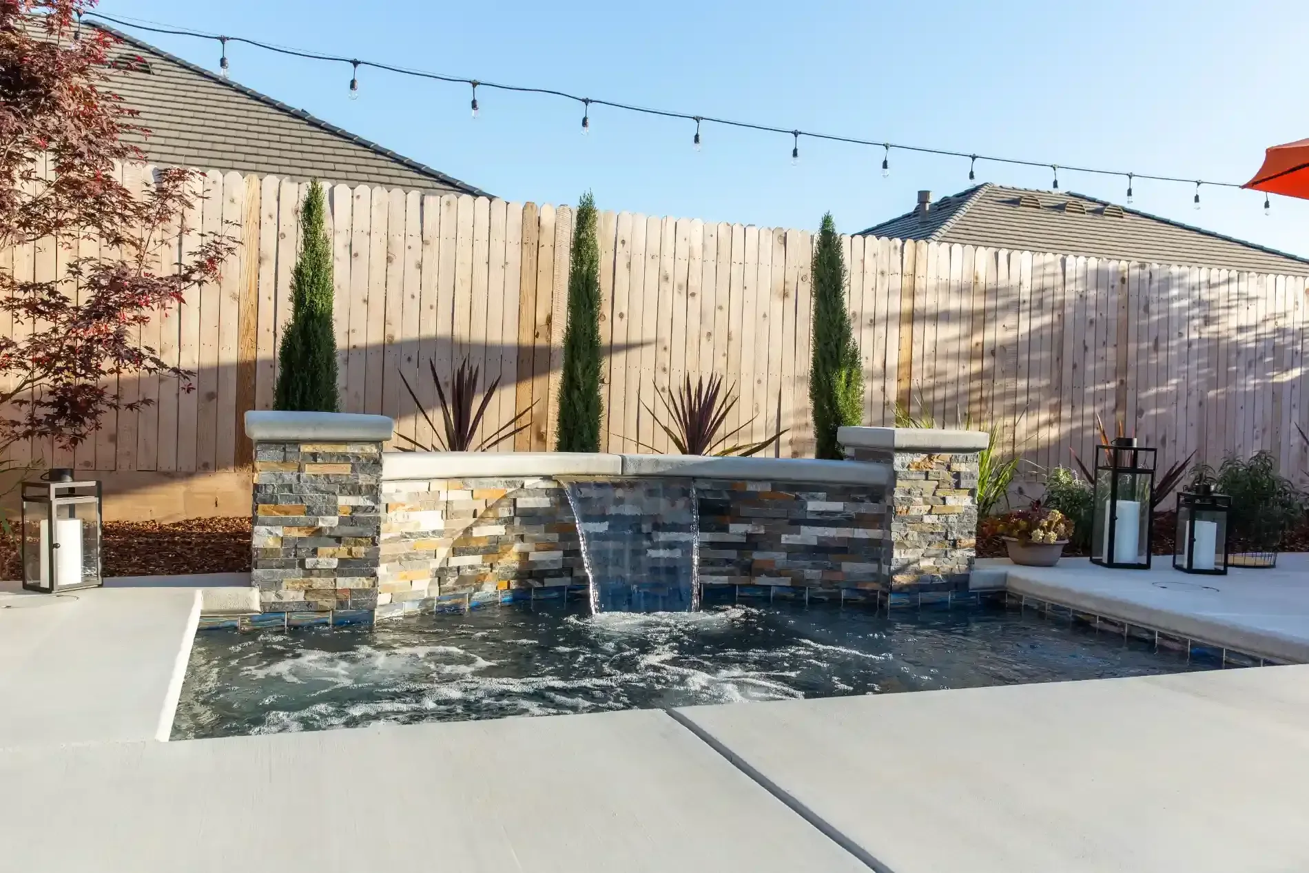 Backyard patio with stone bench, string lights, and potted plants against a wooden fence