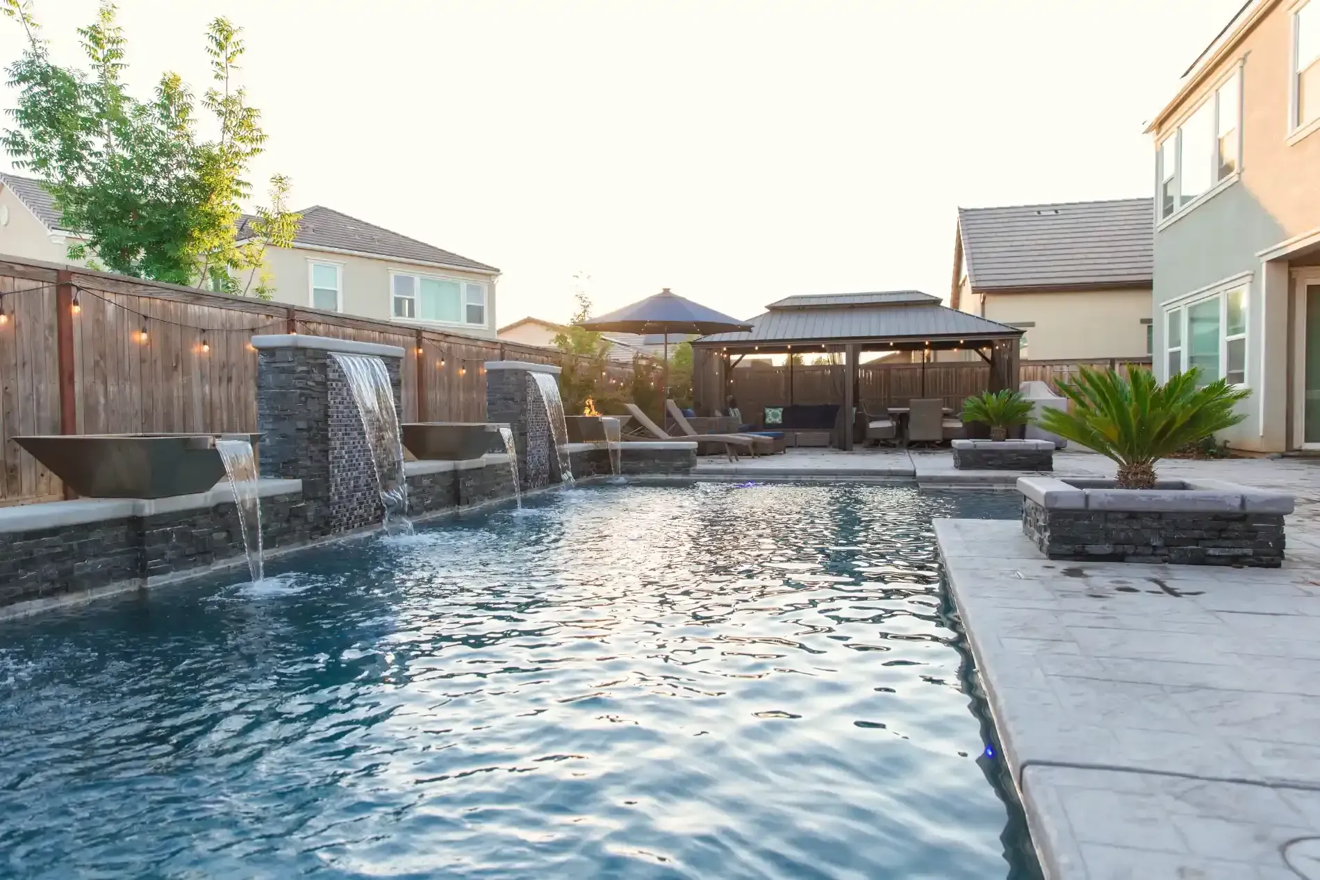 Backyard swimming pool with water features, stone patio, lounge chairs, and covered seating area