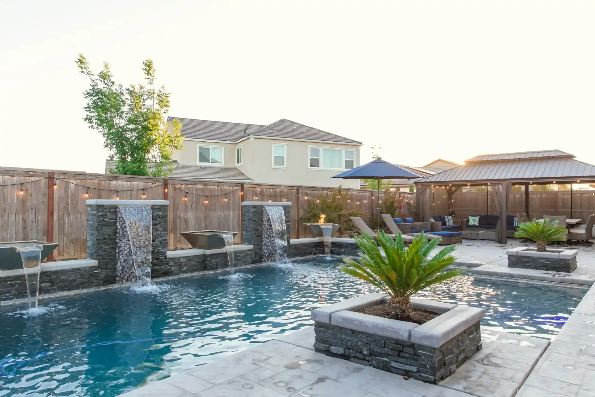 Backyard pool with stone water features, patio seating, and a small palm planter at sunset