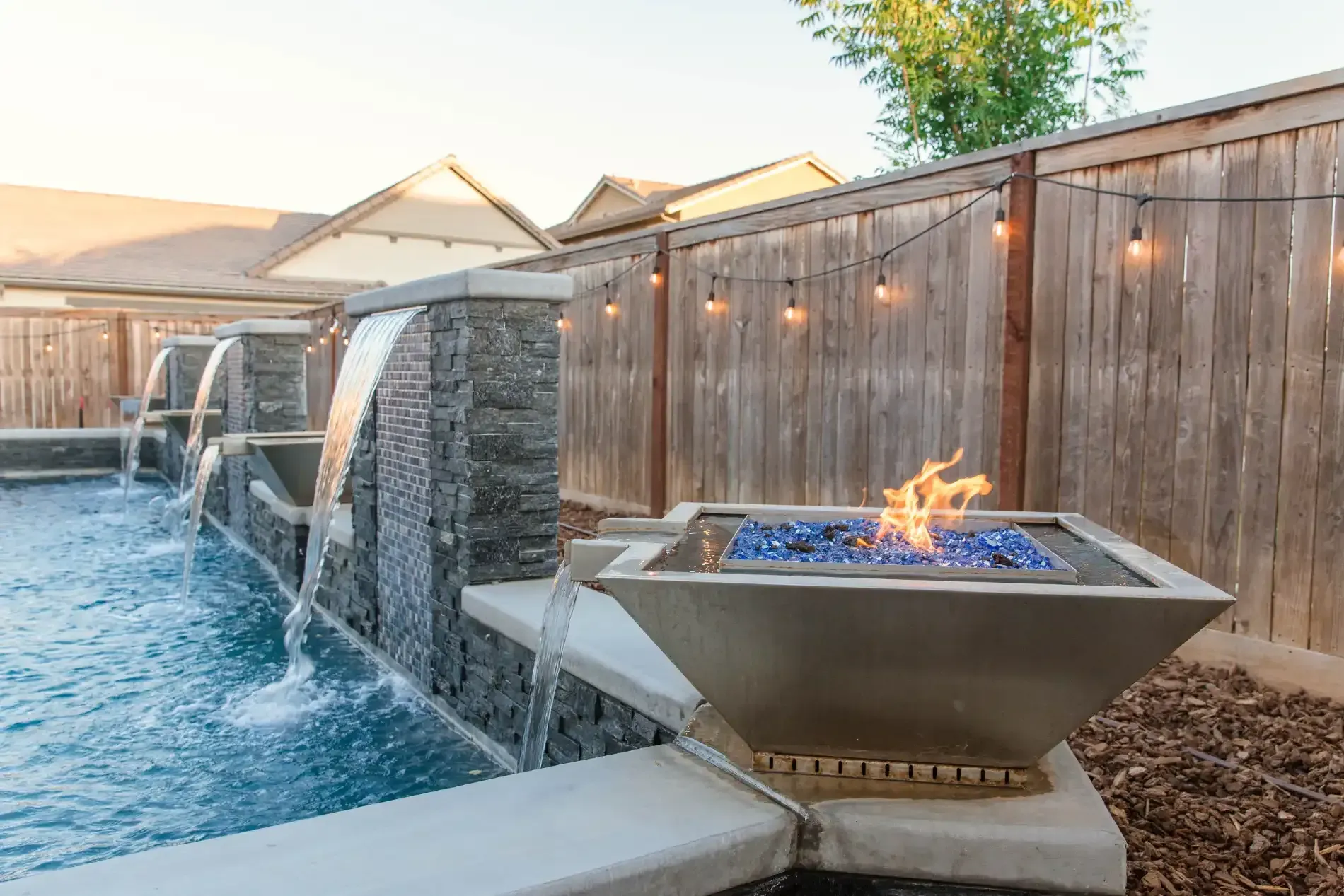 Backyard pool with stone waterfall and lit fire bowl beside a wooden fence