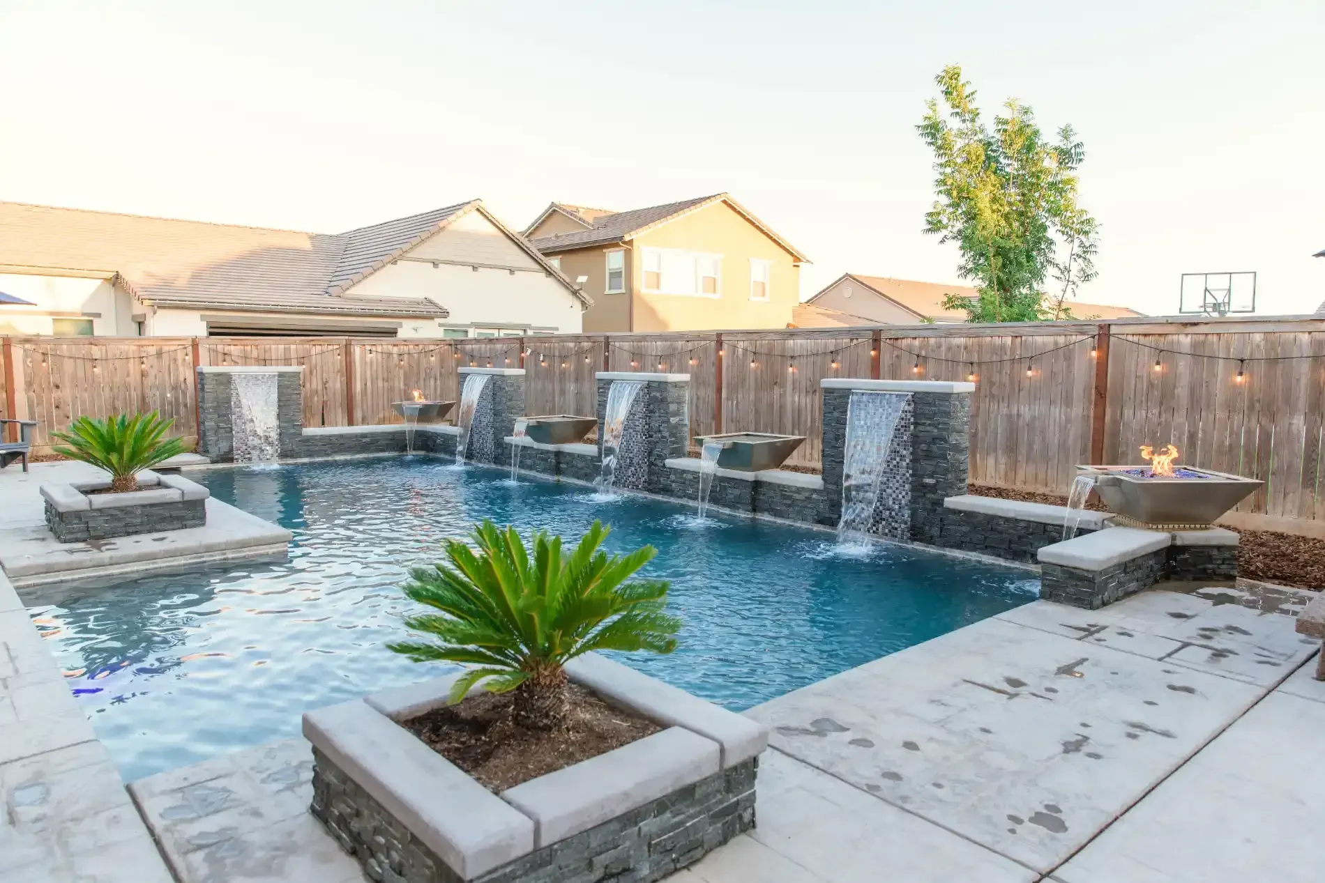 Backyard pool with waterfalls, stone planters, and a basketball hoop at sunset
