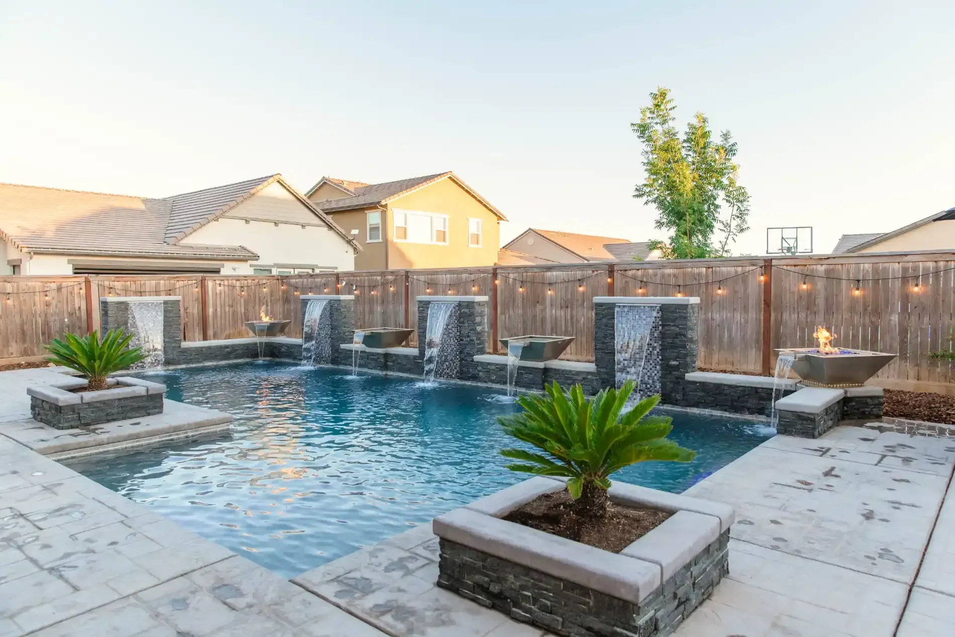 Backyard pool with waterfalls, stone deck, and potted plants at sunset