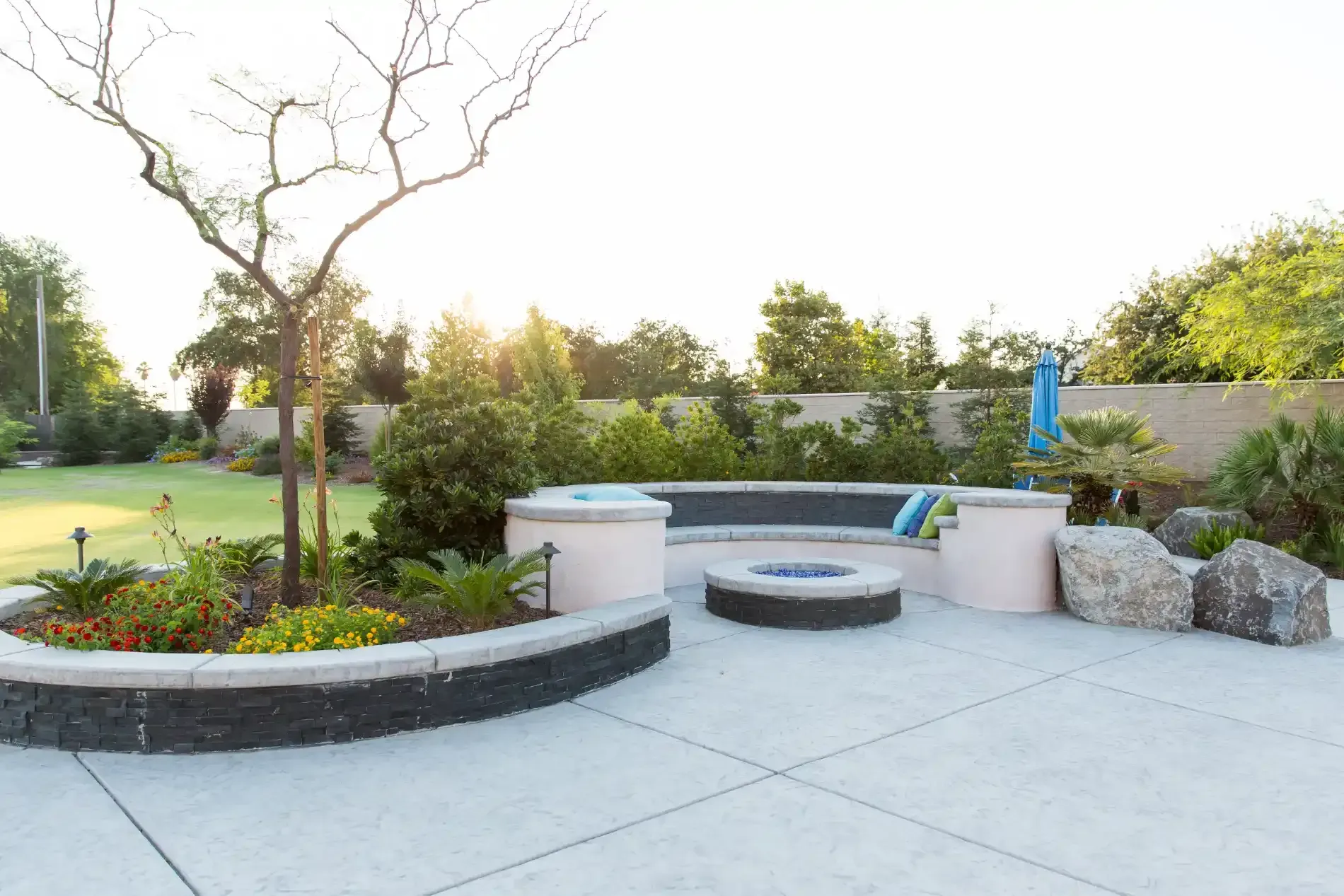 Modern backyard patio with curved stone seating, fire pit, landscaping, and a blue umbrella